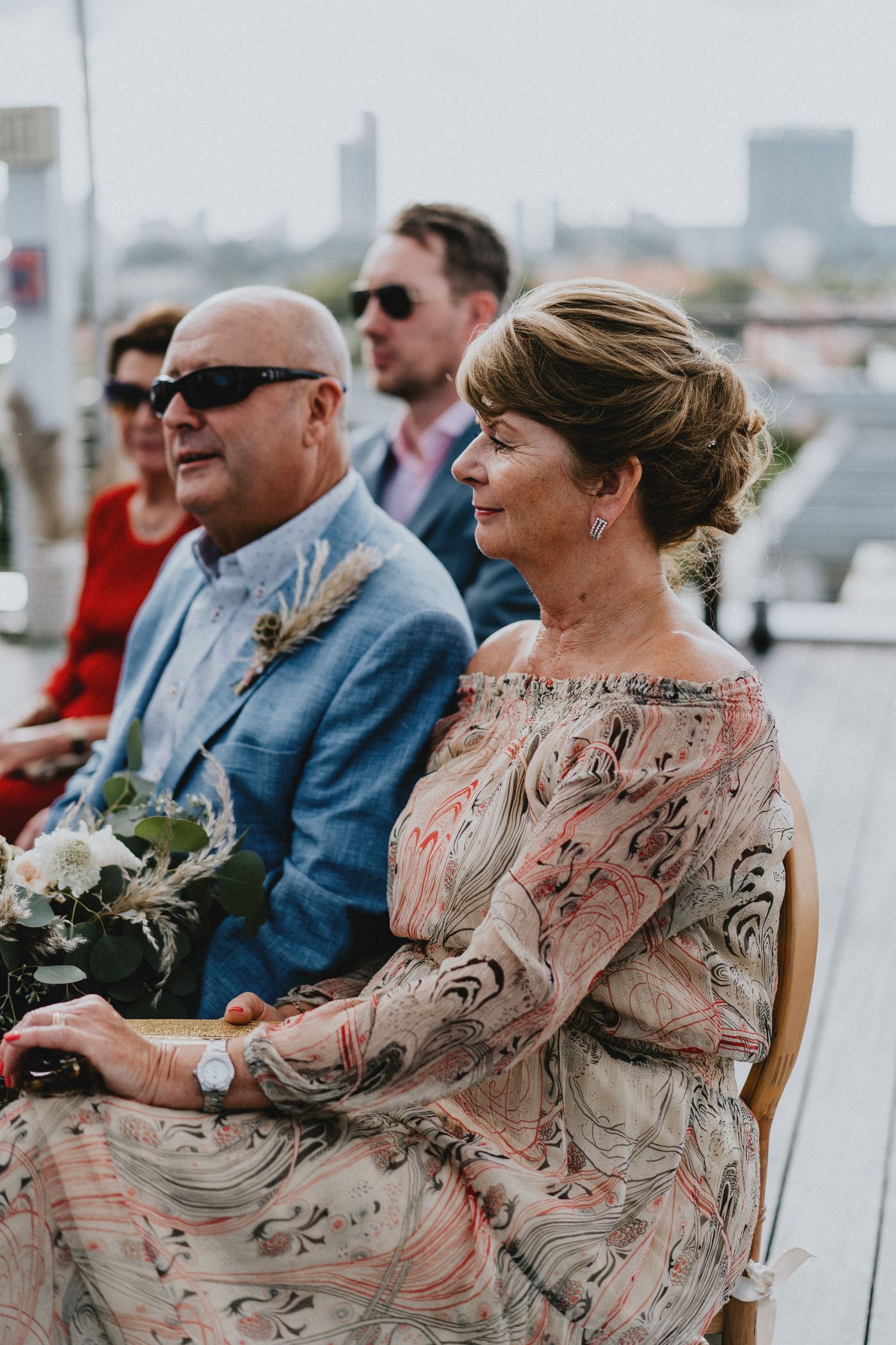 Elegant wedding guests seated outdoors, woman in floral off-shoulder dress beside man in blue suit with sunglasses.