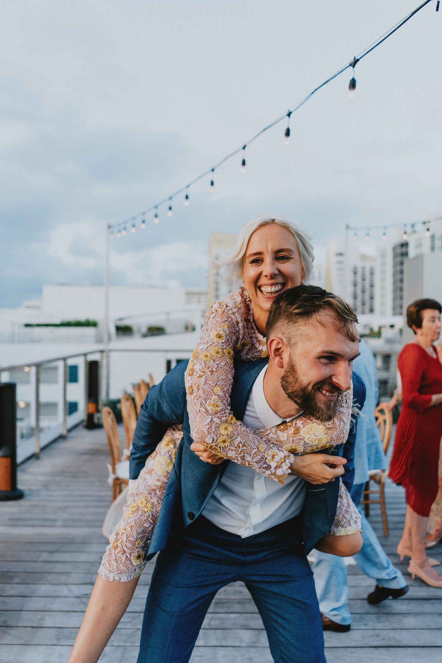 A joyful groom gives his bride a piggyback ride on a rooftop deck with string lights at their outdoor wedding reception.