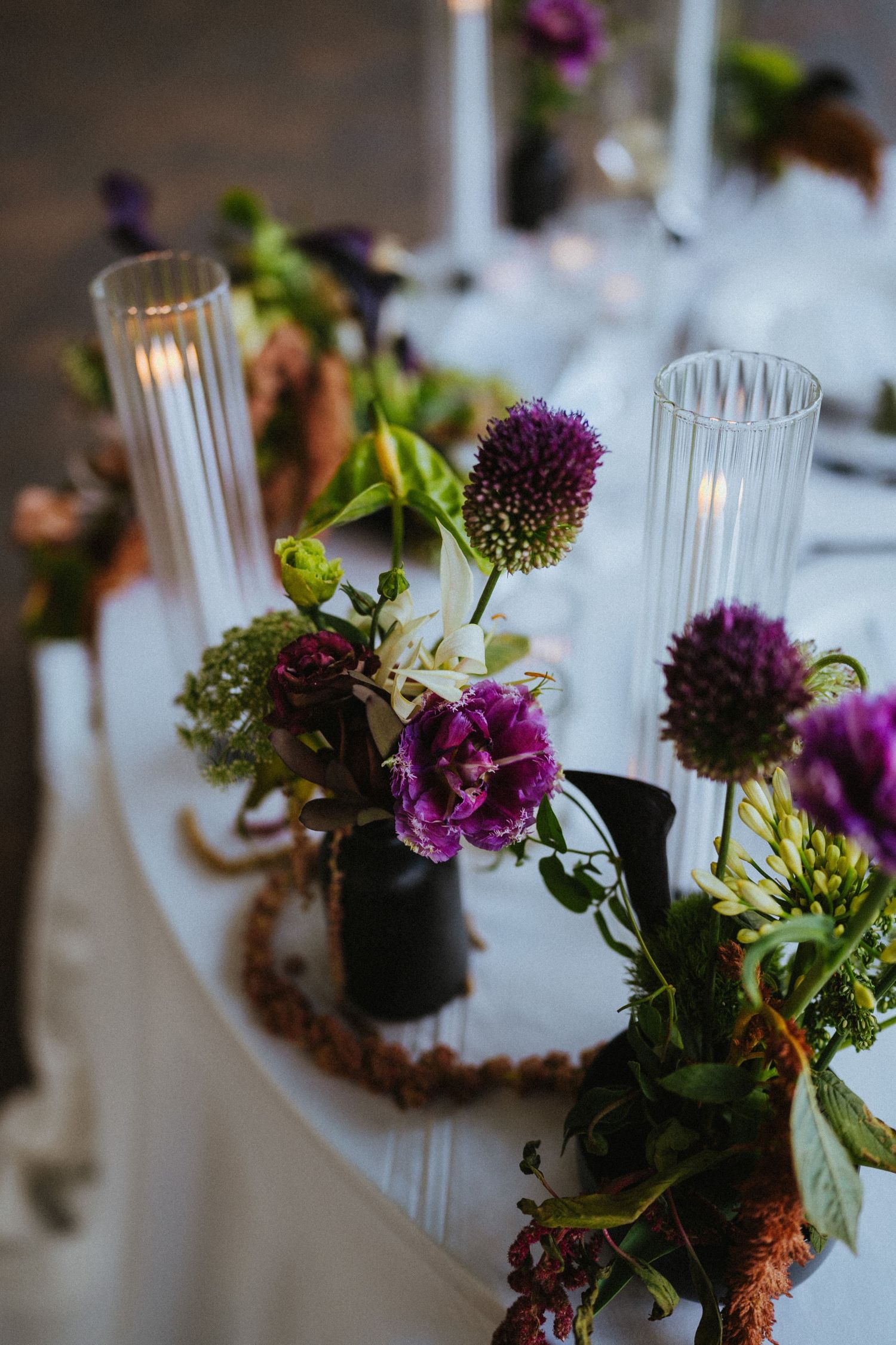 Elegant wedding table centerpiece with purple allium flowers, dark vases, and glass candle holders on a white tablecloth.