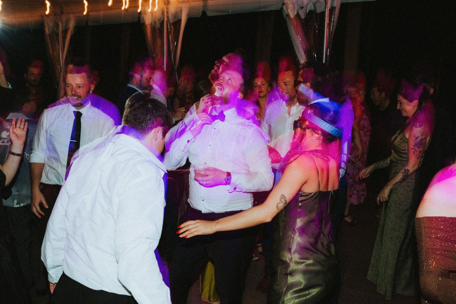 Guests dancing energetically at a wedding reception under colorful purple and pink lights.