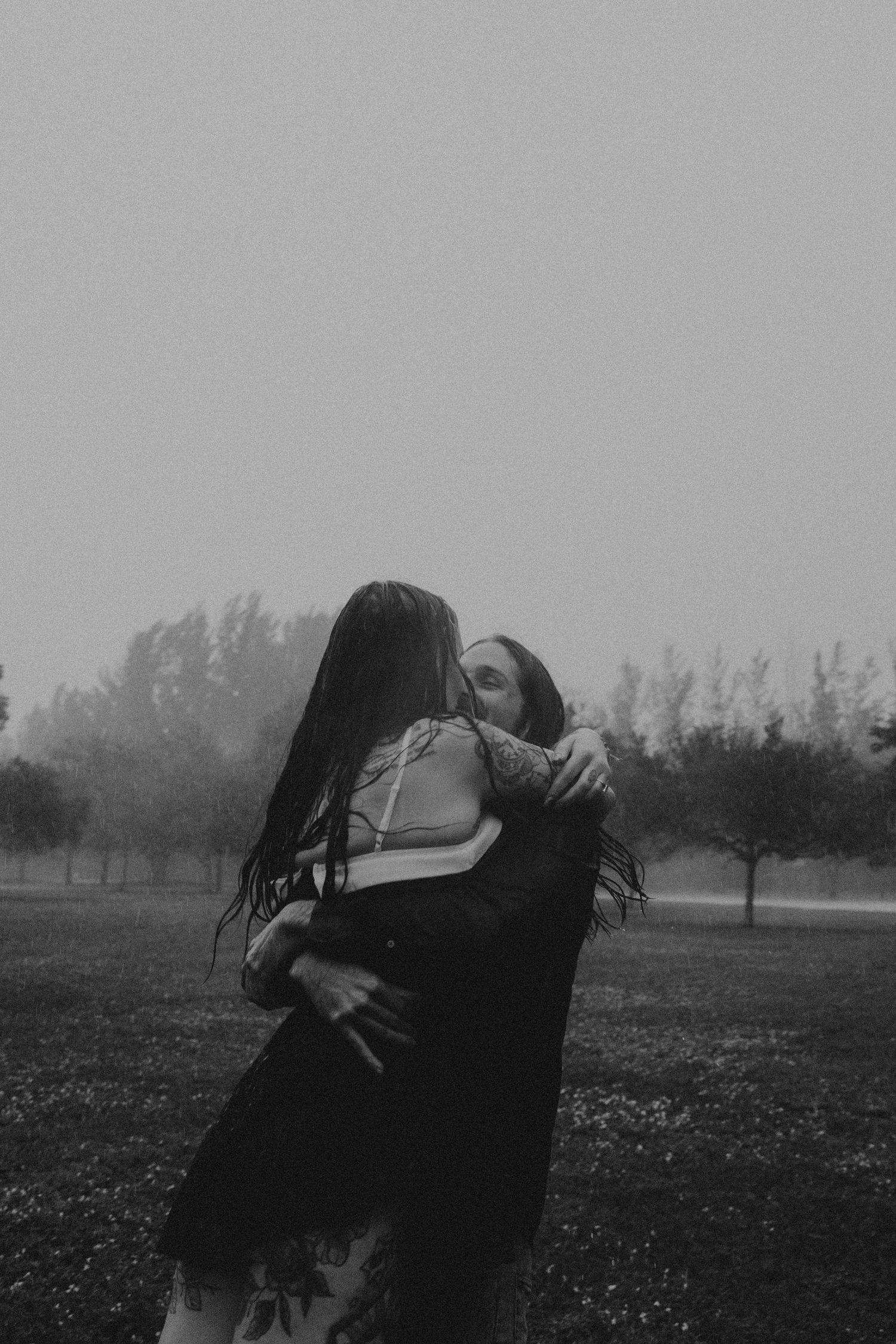 A tender embrace between two people in a misty field with trees silhouetted in the background, captured in black and white.