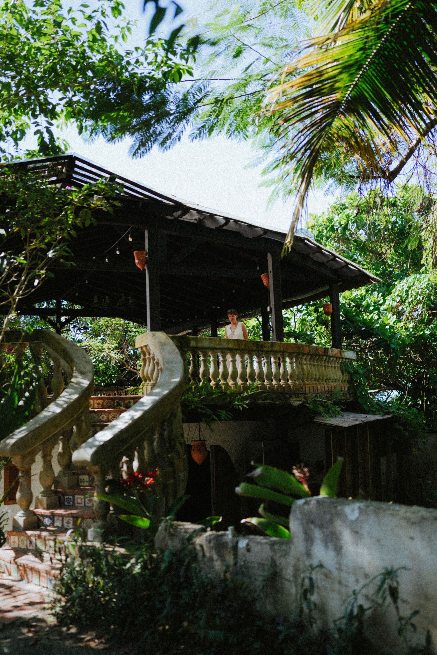 Tropical garden pavilion with ornate white balustrade, lush palm trees, and a person standing under a dark metal roof.