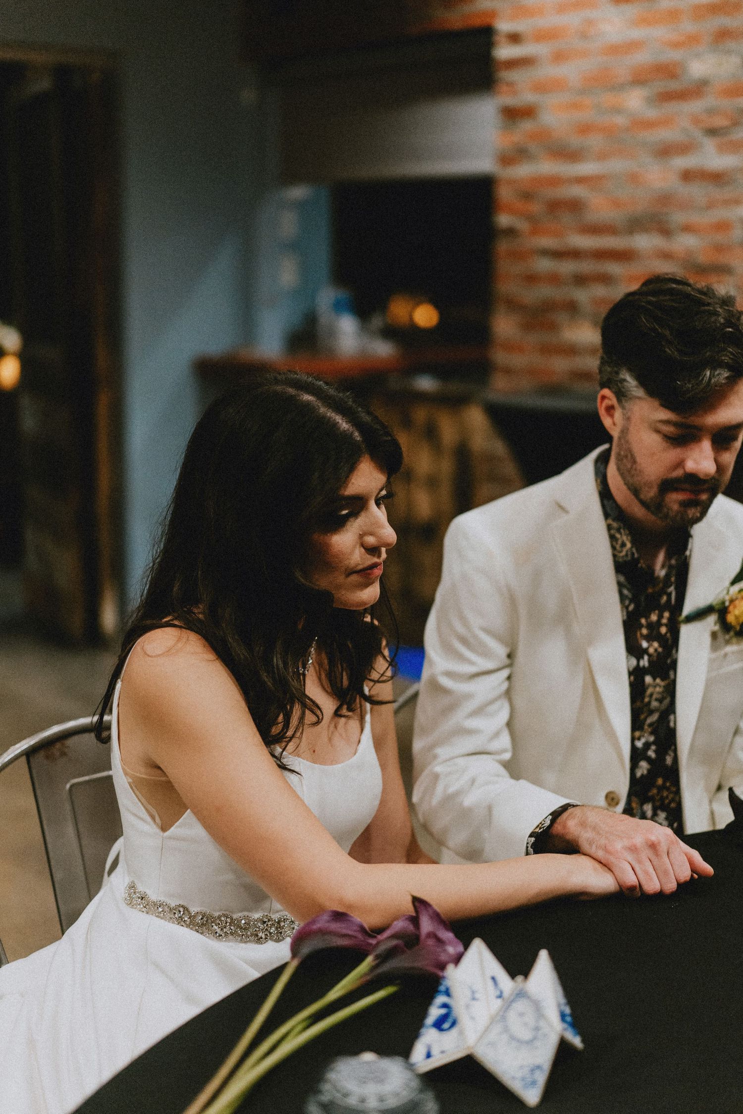 Bride in white gown and groom in white blazer sit together at wedding reception table with purple flowers.