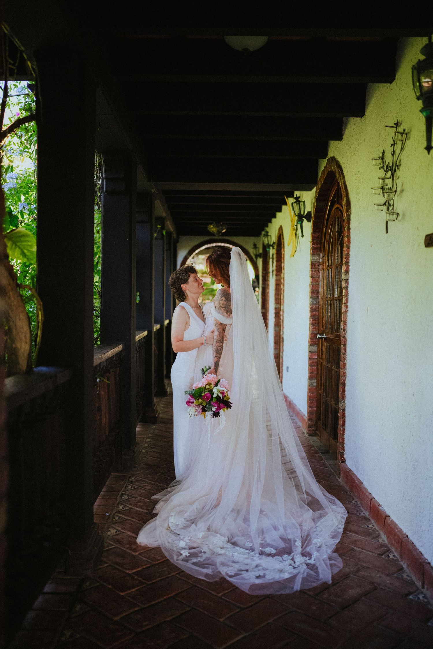 Two brides share a romantic kiss in a sunlit corridor, one wearing a flowing white gown with a long lace veil and holding a bouquet.