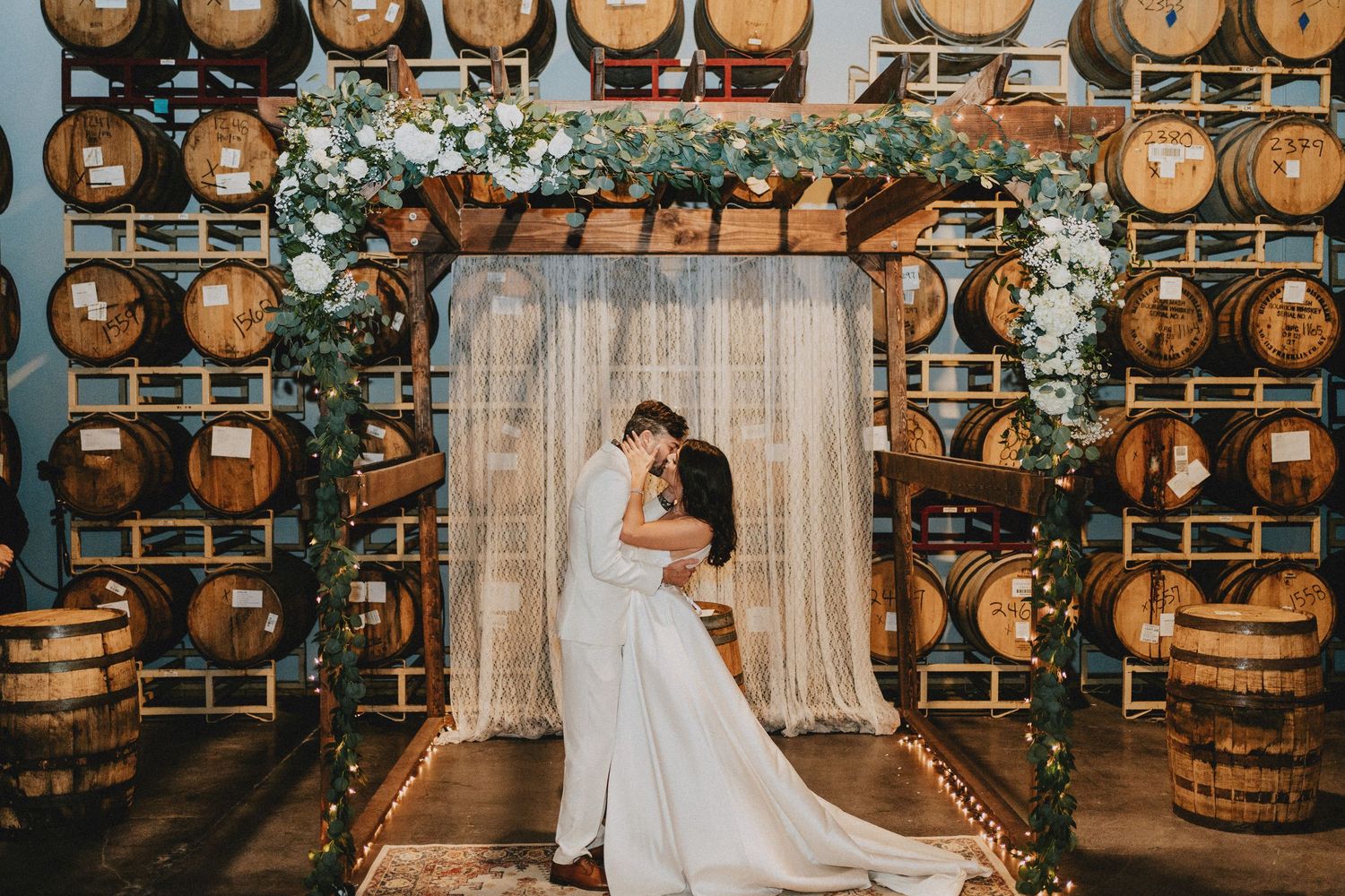Couple sharing first kiss under floral arch with greenery, surrounded by wooden wine barrels at winery wedding venue.