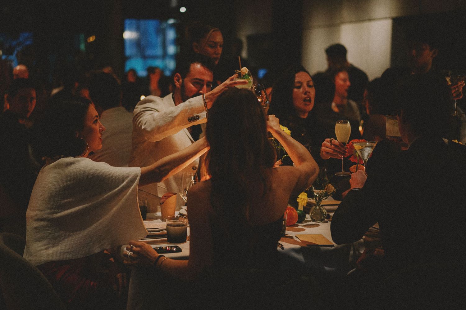 Group of people raising glasses in a toast at a dimly lit dinner party or wedding reception.
