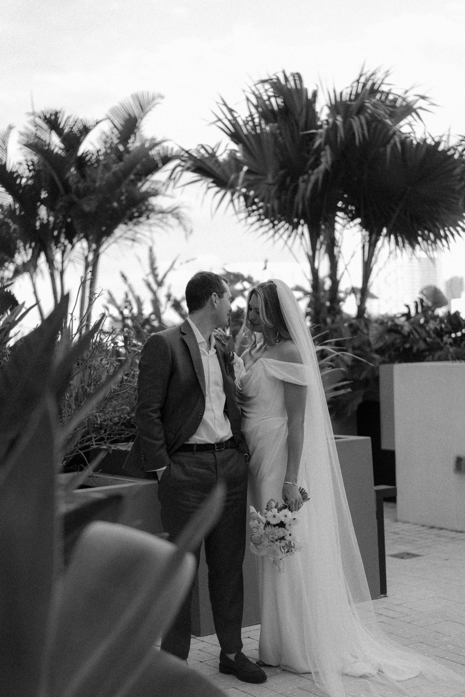 Black and white photo of a bride and groom sharing a romantic moment outdoors, surrounded by tropical palm trees.