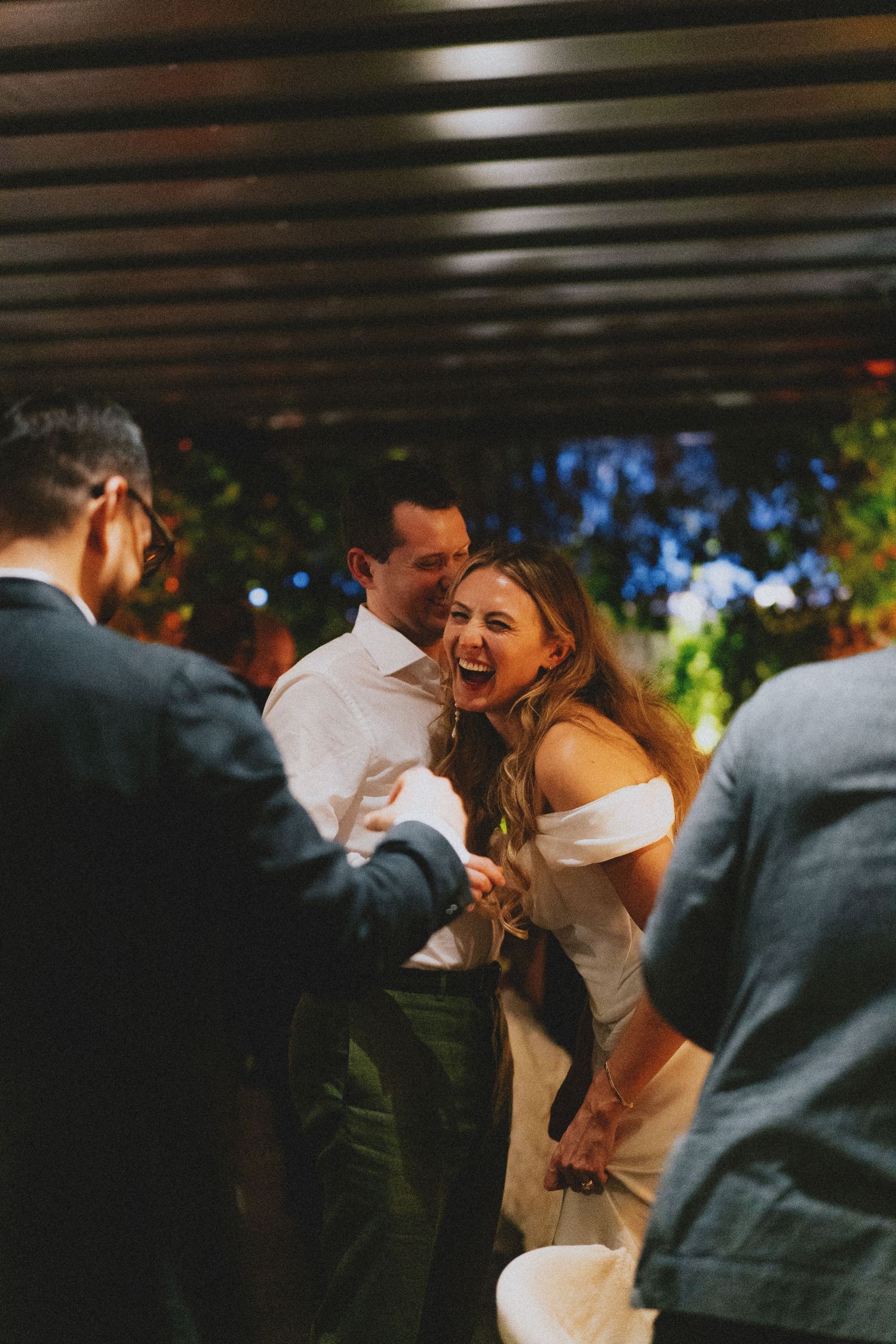 Joyful bride in off-shoulder gown laughing with guests during outdoor wedding reception under pergola.