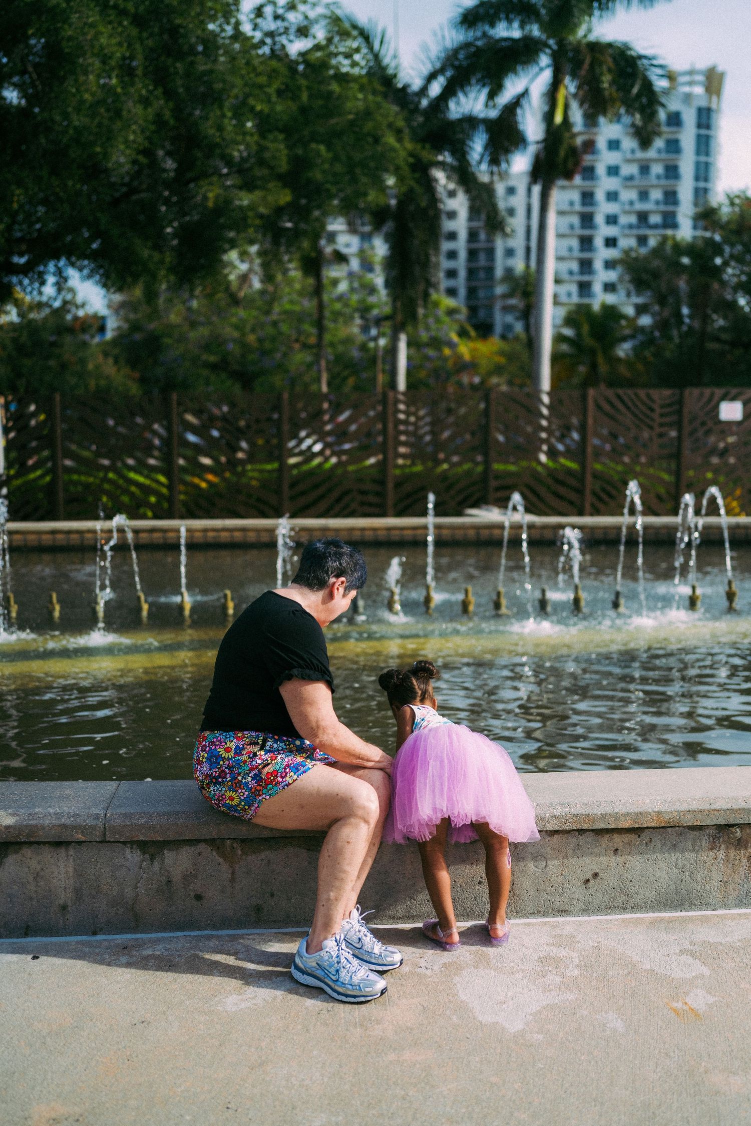 Adult and toddler girl in pink tutu playing by a decorative fountain in a sunny urban park with palm trees.