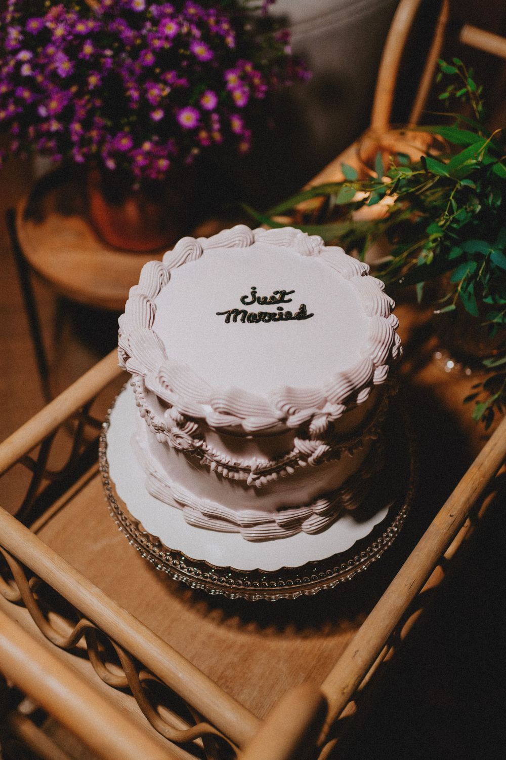 White wedding cake with 'Just Married' text displayed on wooden stand with purple flowers in background.
