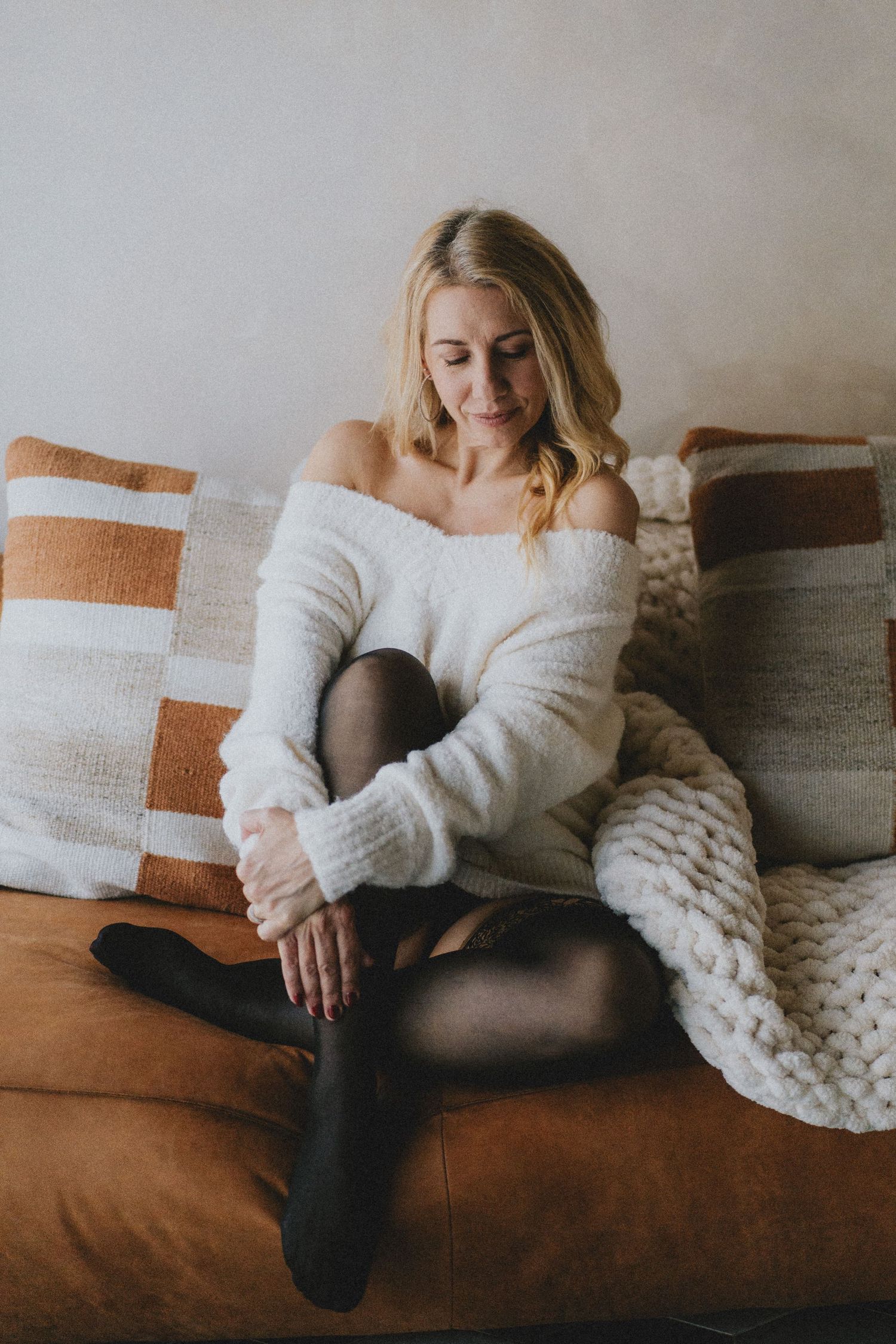 Cozy lifestyle portrait of someone relaxing on brown couch wearing white sweater, black tights and chunky knit blanket.