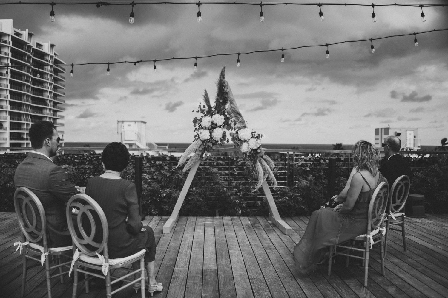 Black and white rooftop wedding ceremony with floral arch, string lights, and guests seated on elegant chairs overlooking cityscape.