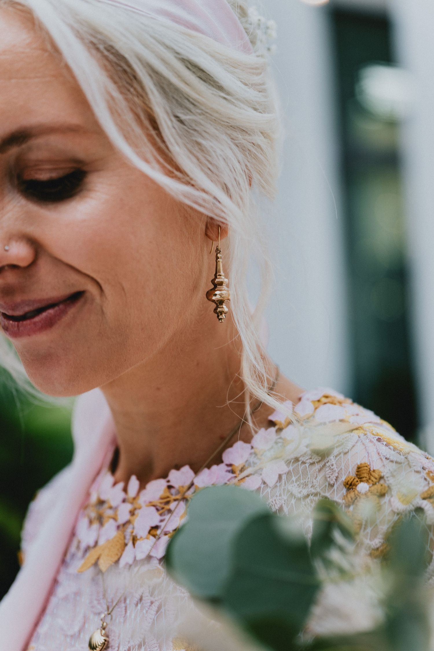 Close-up of a smiling blonde bride wearing gold drop earrings and a floral embroidered dress, holding a bouquet.