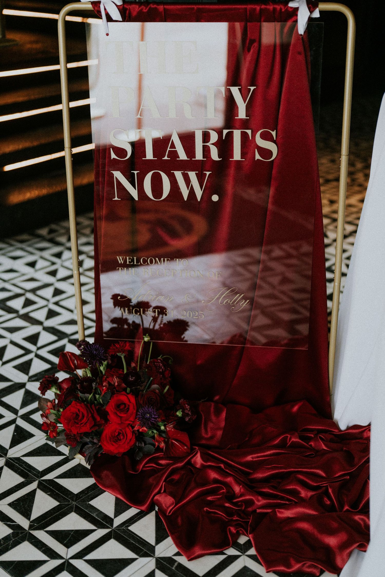 Acrylic wedding welcome sign reading The Party Starts Now with red roses and burgundy satin draping.