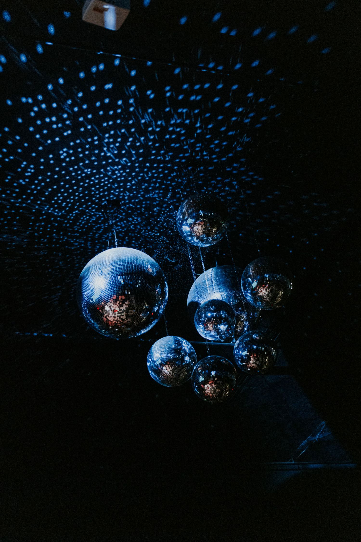 Cluster of mirrored disco balls hanging from a dark ceiling with blue light reflections creating a dazzling pattern.