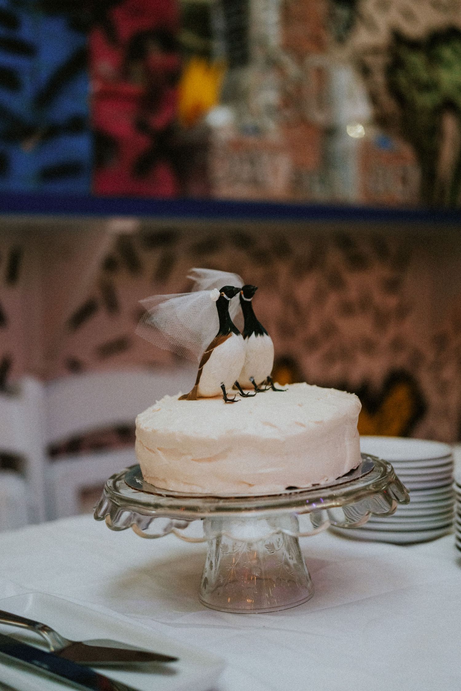 White wedding cake on glass stand topped with bird figurines wearing a bridal veil.