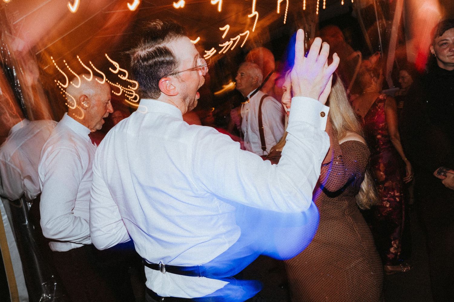 Wedding guest dancing energetically under warm string lights at a lively indoor reception celebration.