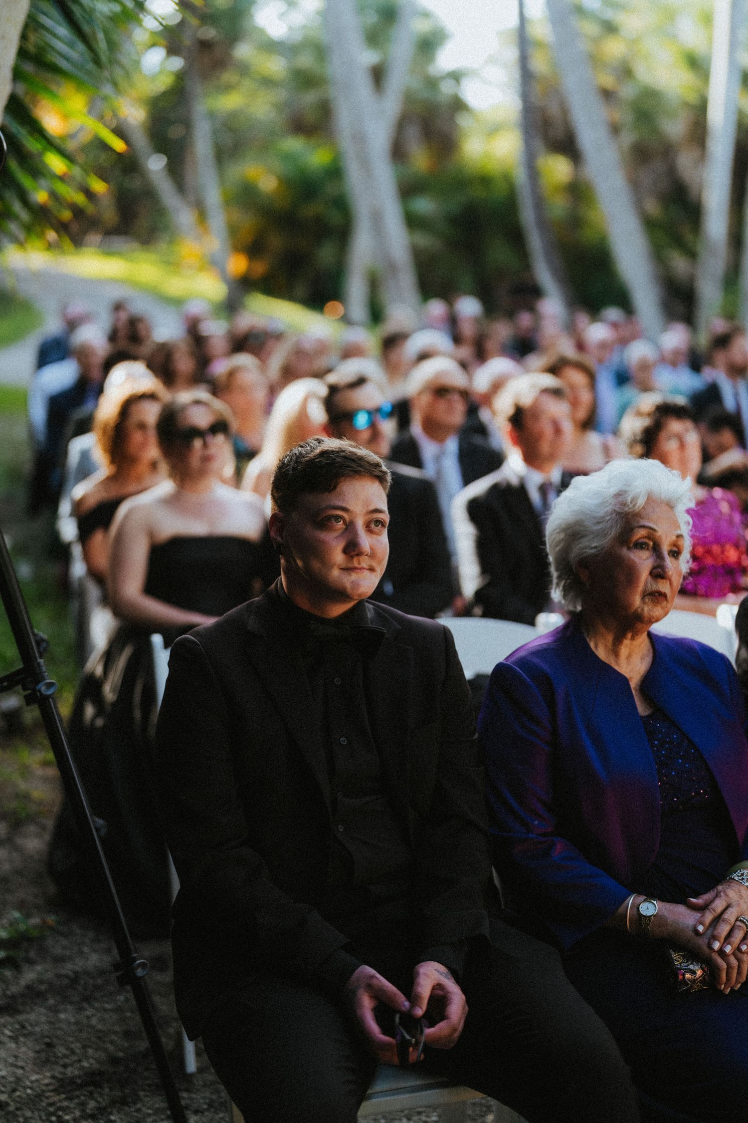 Wedding guests seated outdoors among palm trees watch emotional ceremony in natural garden setting.