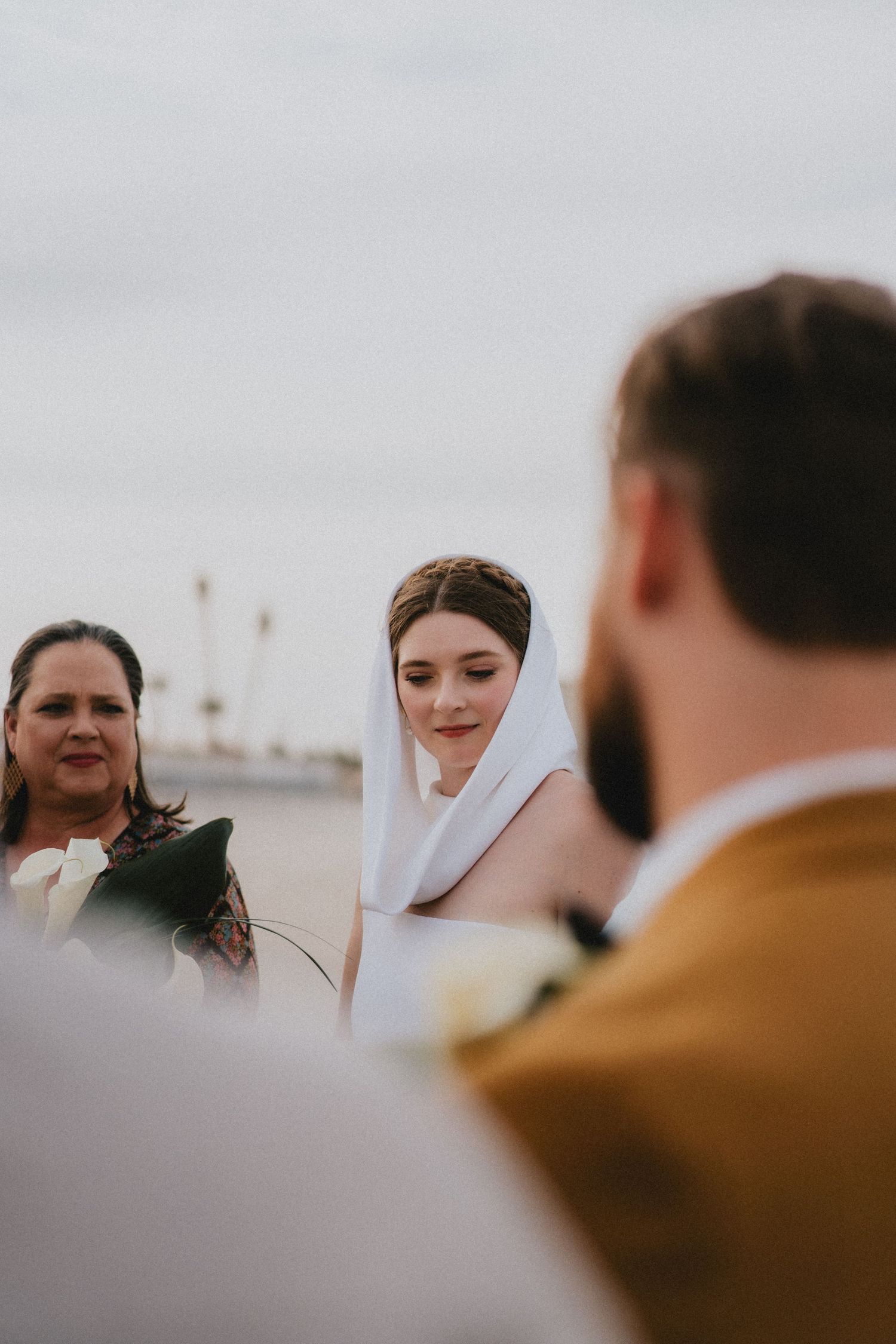 Bride in white veil gazes down during emotional outdoor beach wedding ceremony.