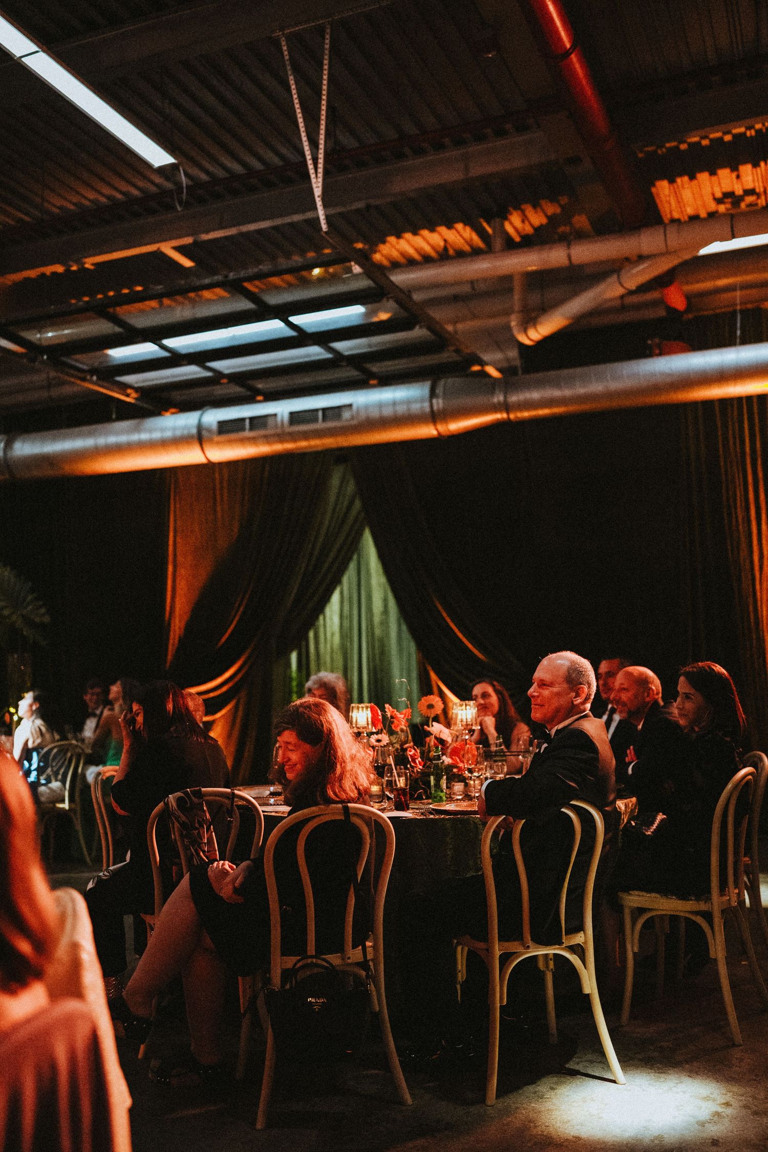 Guests seated at round tables in a dimly lit industrial venue with dark curtains and warm lighting.