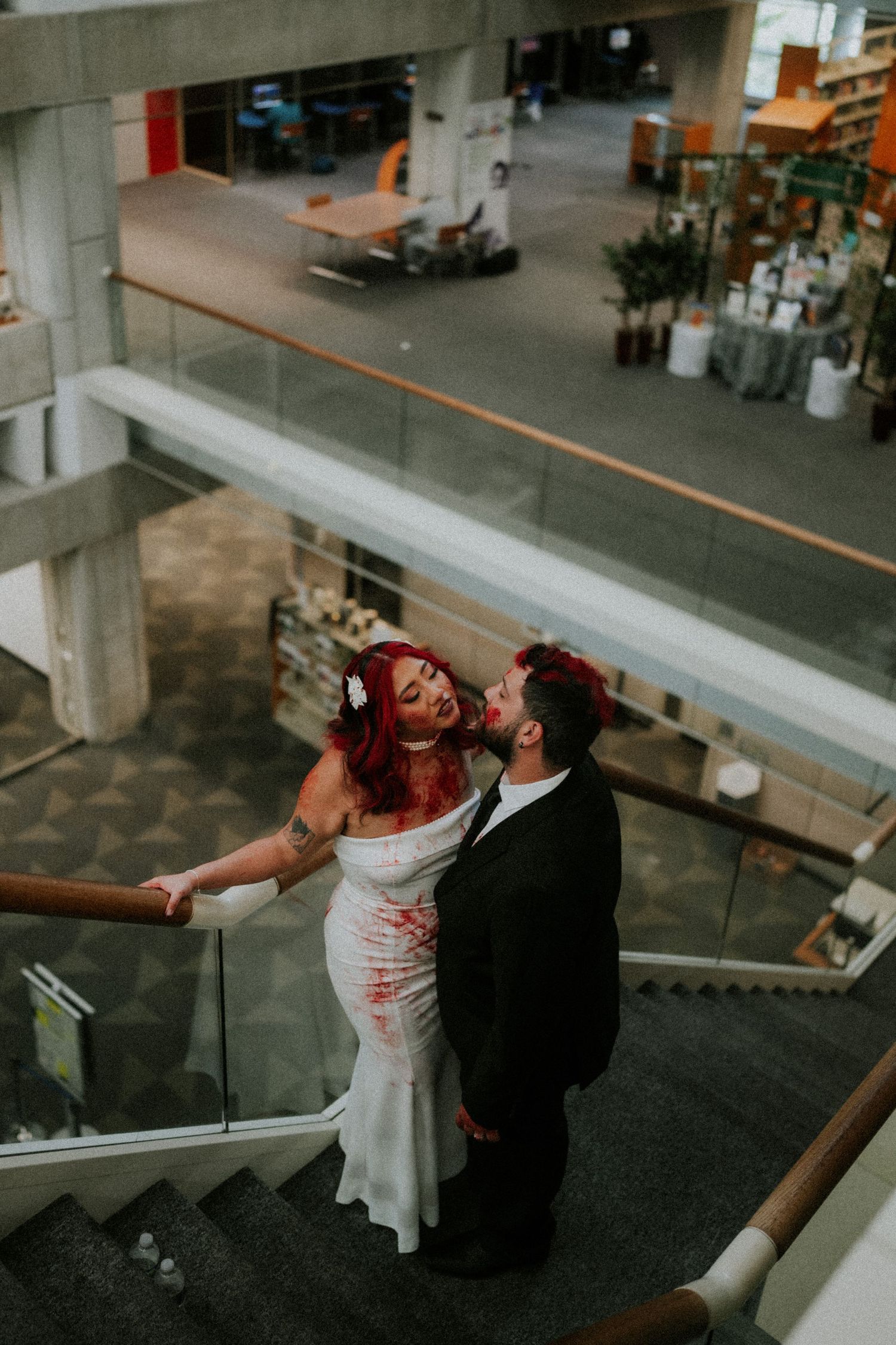 Couple in zombie wedding attire with fake blood pose romantically on stairs inside a modern library.