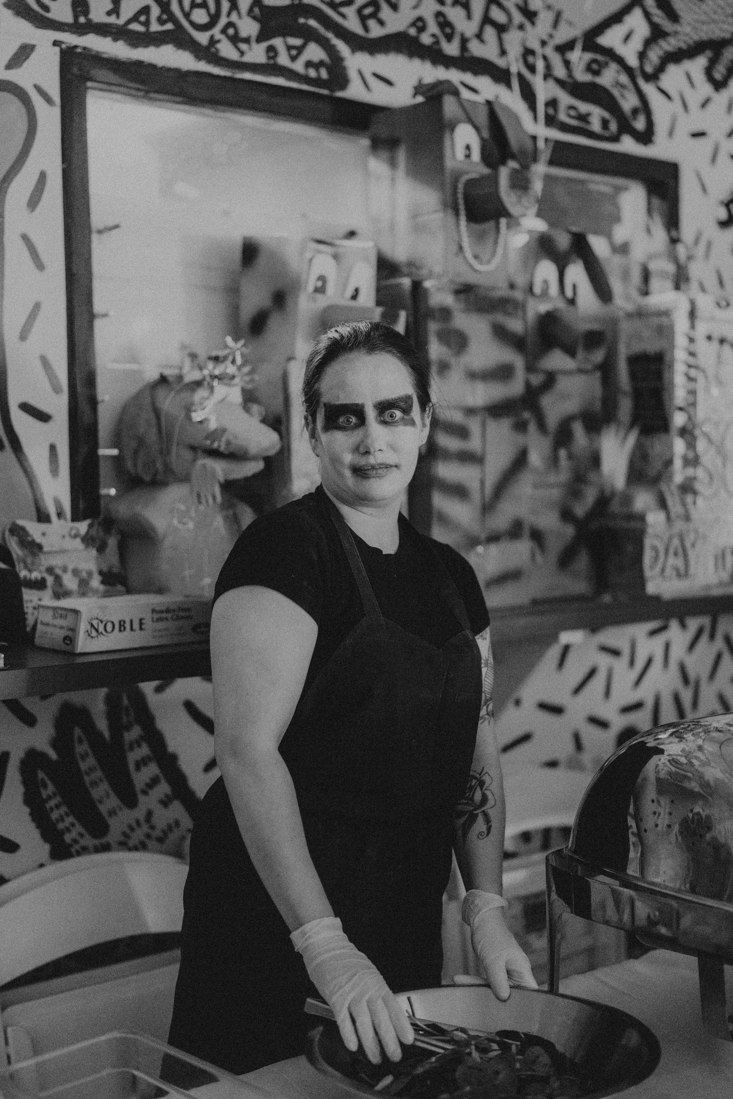Black and white photo of a cook in Halloween makeup and apron working in a graffiti-decorated kitchen.