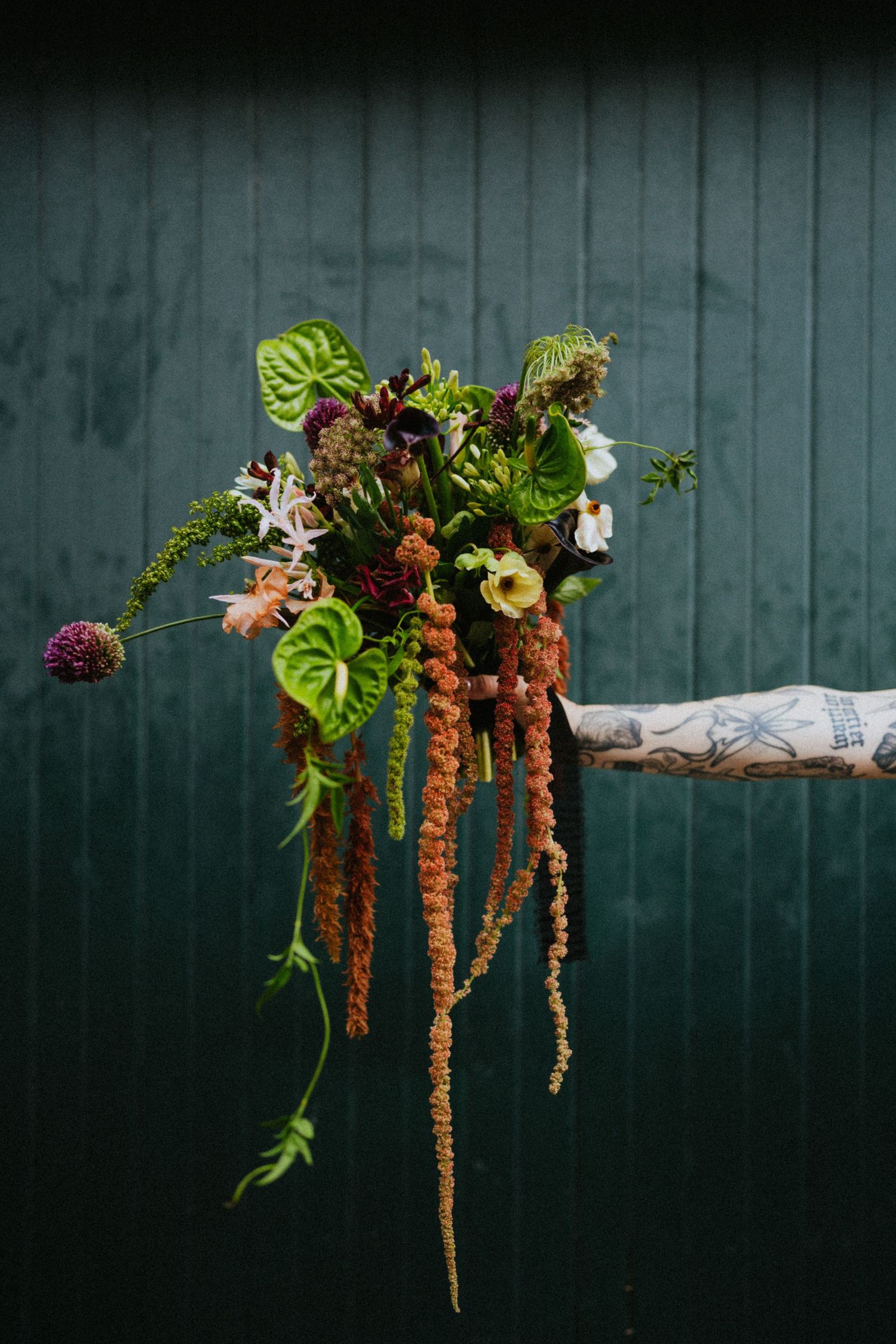 Tattooed hand holding a wild botanical bouquet with hanging amaranthus, anthurium, and allium against a dark green backdrop.