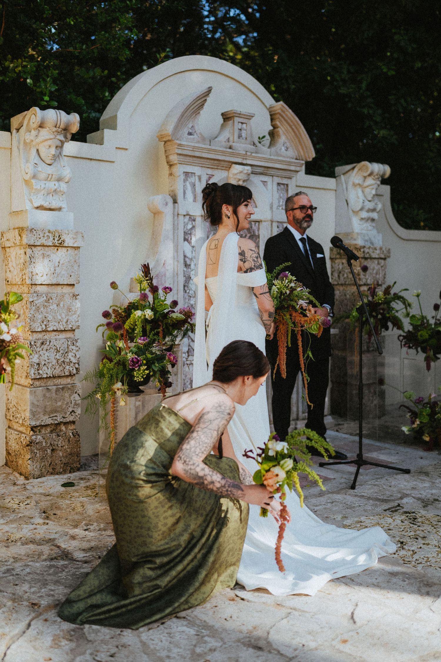 Bridesmaid in gold dress adjusts bride's train during outdoor garden wedding ceremony with floral arrangements.