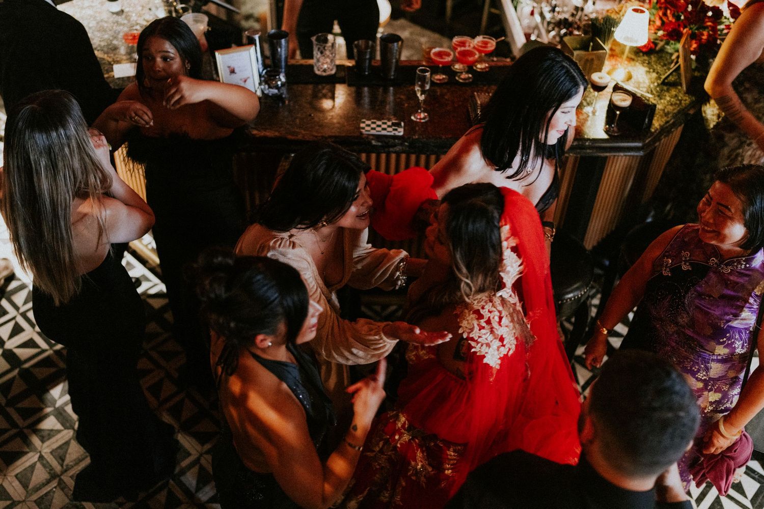 Guests gather around in a lively wedding reception scene, with a woman in a red dress at the center.