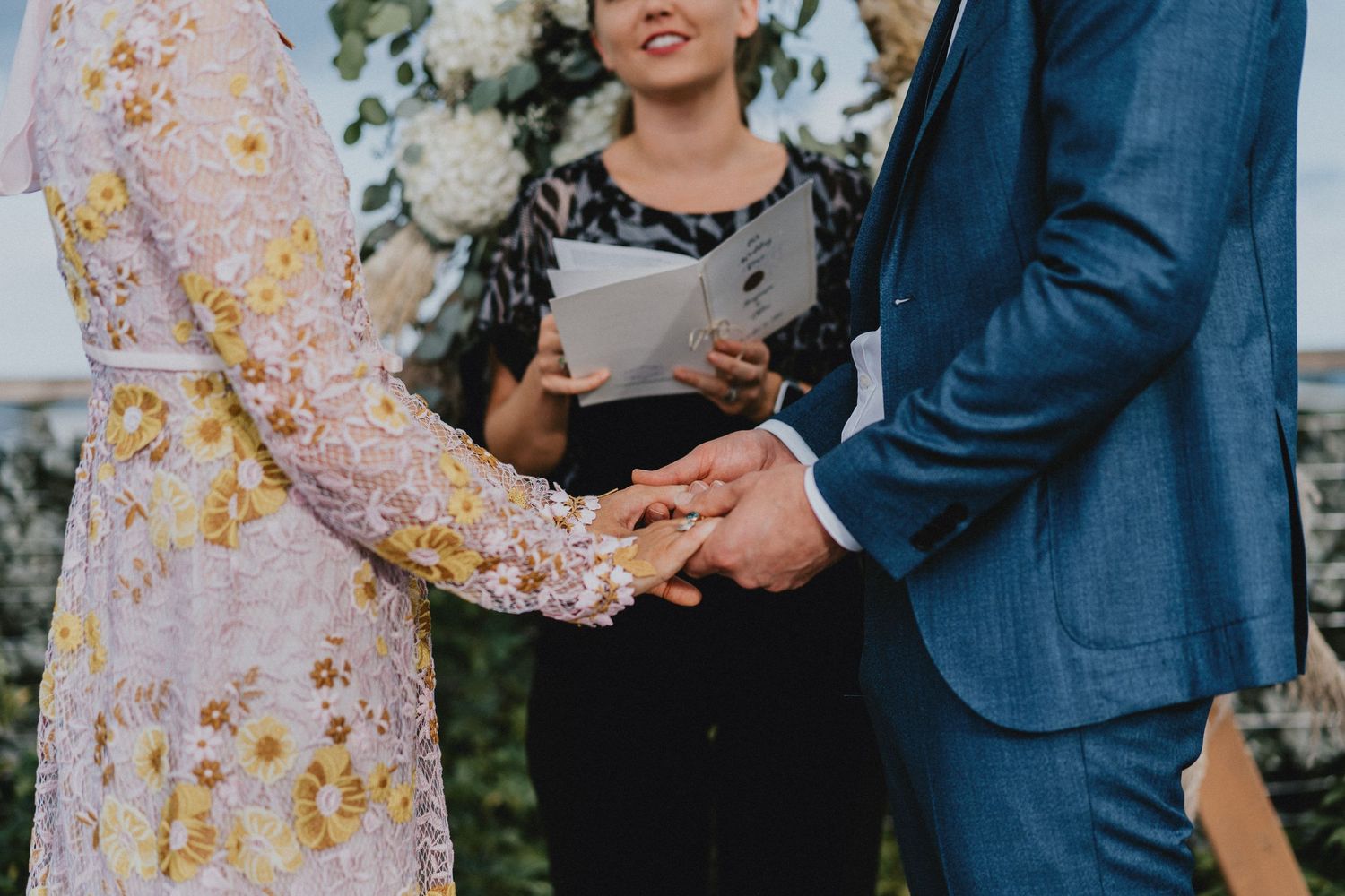 Couple exchanging rings at outdoor wedding ceremony with officiant holding book in background.
