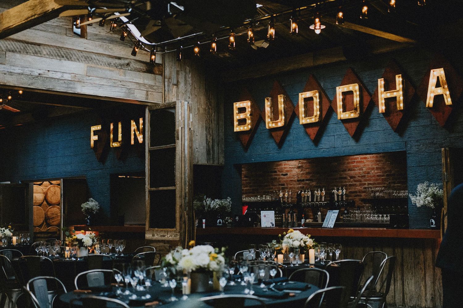 Rustic wedding venue bar with Buddha and Fun marquee signs, string lights, and elegant table settings.