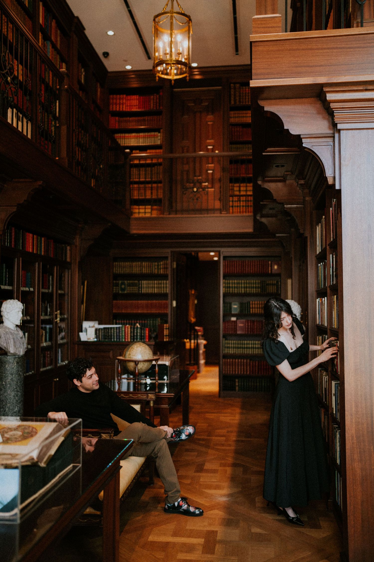 Couple in a grand vintage library with dark wood shelves filled with books, woman in black dress, man seated on floor.