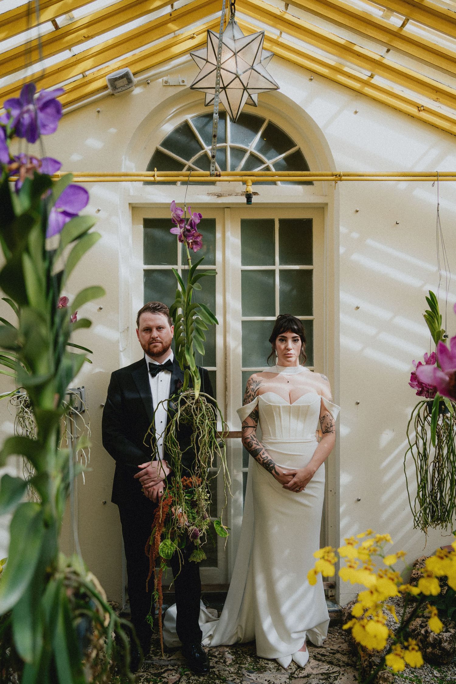 Couple in wedding attire poses in a greenhouse with orchids, groom in tuxedo, bride in white gown with tattoos.