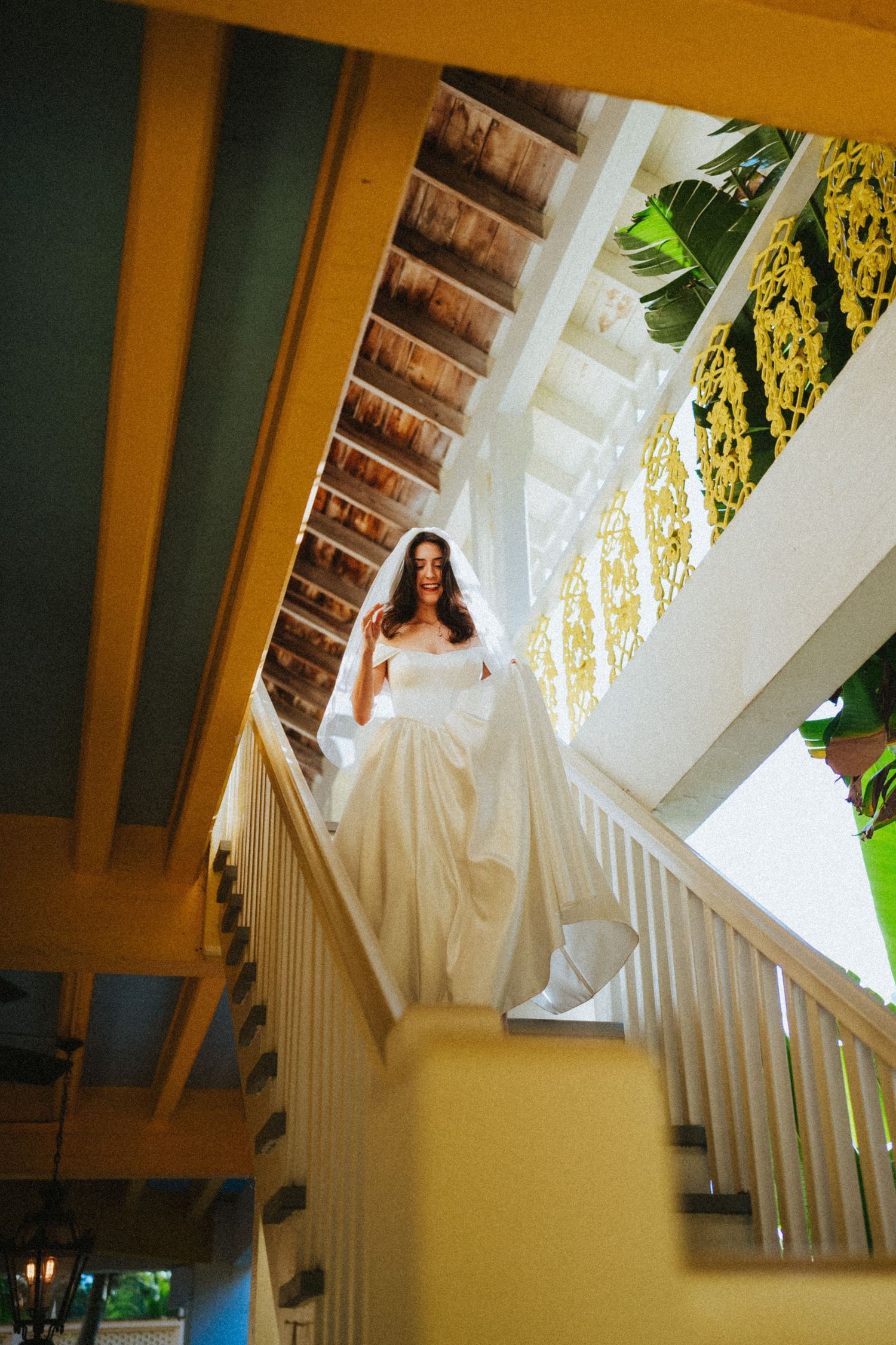 Bride in white wedding gown and veil descends elegant staircase with white railings and tropical greenery.