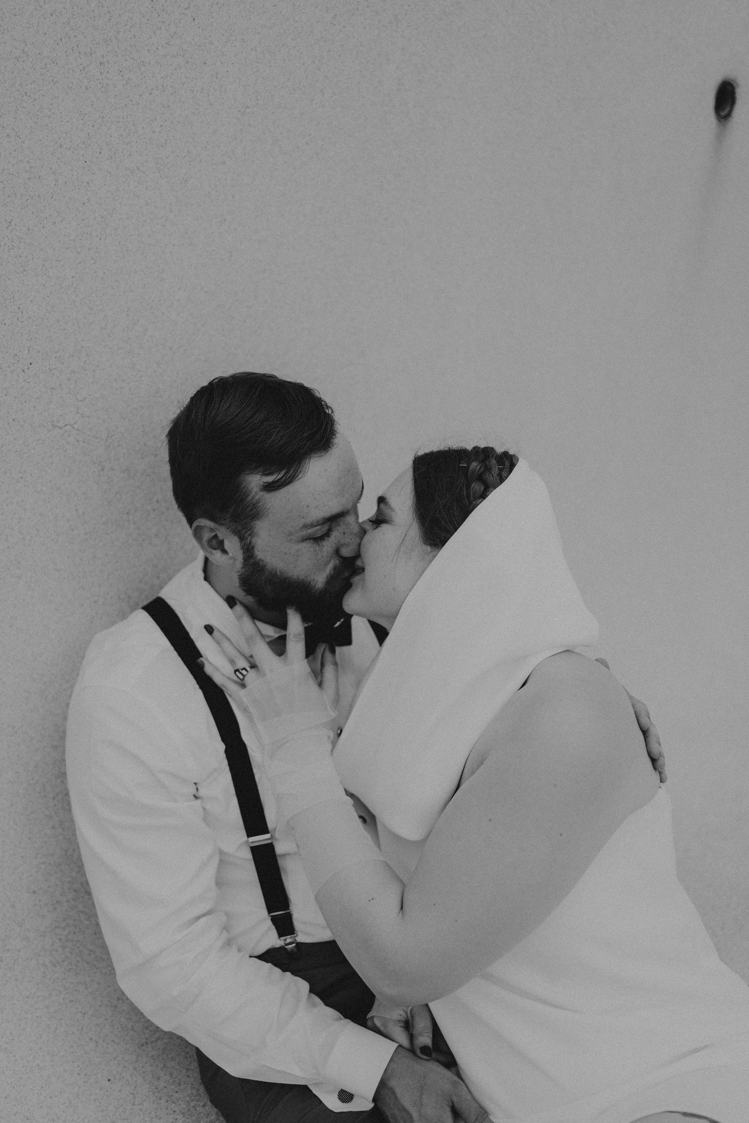 Black and white photo of bride and groom sharing a tender kiss, groom wearing suspenders and bow tie.