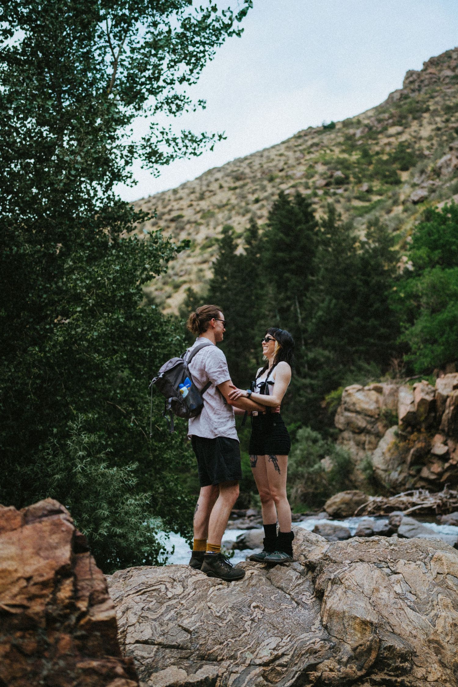 Couple holding hands on a rocky mountain overlook with a river and green trees behind them.