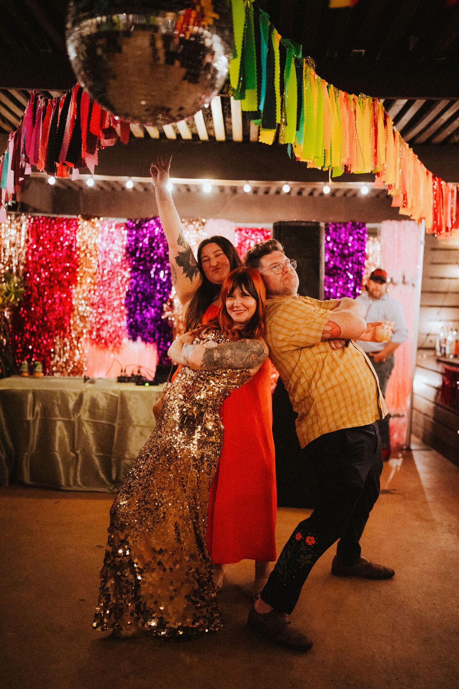 Three friends posing and dancing under a disco ball with rainbow fringe decorations at a festive party.