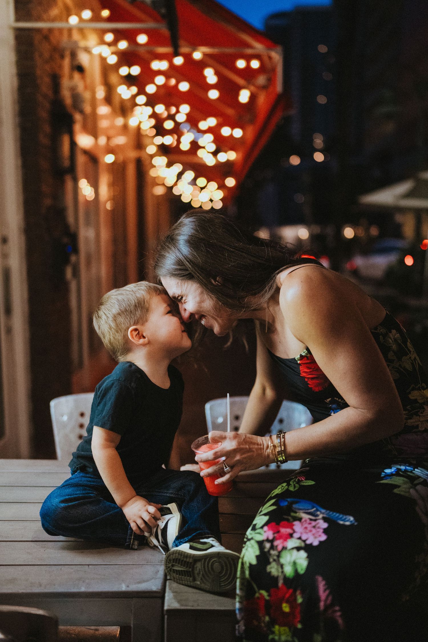 A tender moment shared between parent and child on a porch at dusk with twinkling string lights in the background.