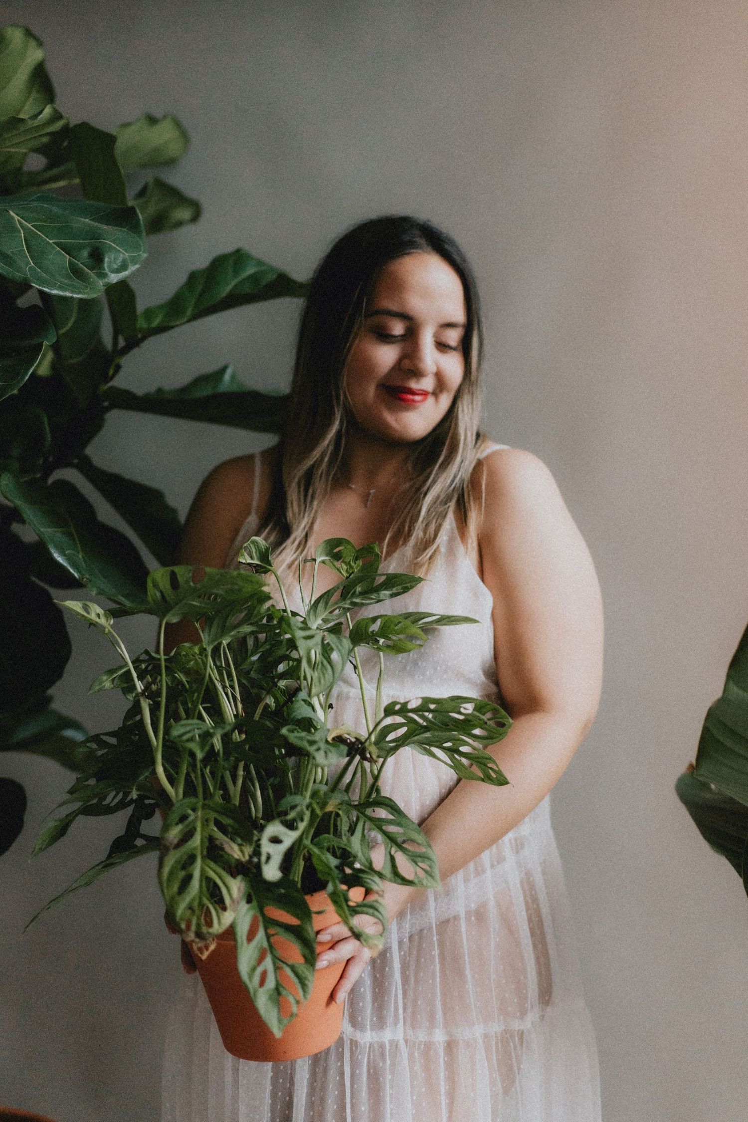 Woman in white dress holds and admires potted monstera plant against neutral wall in soft natural lighting.