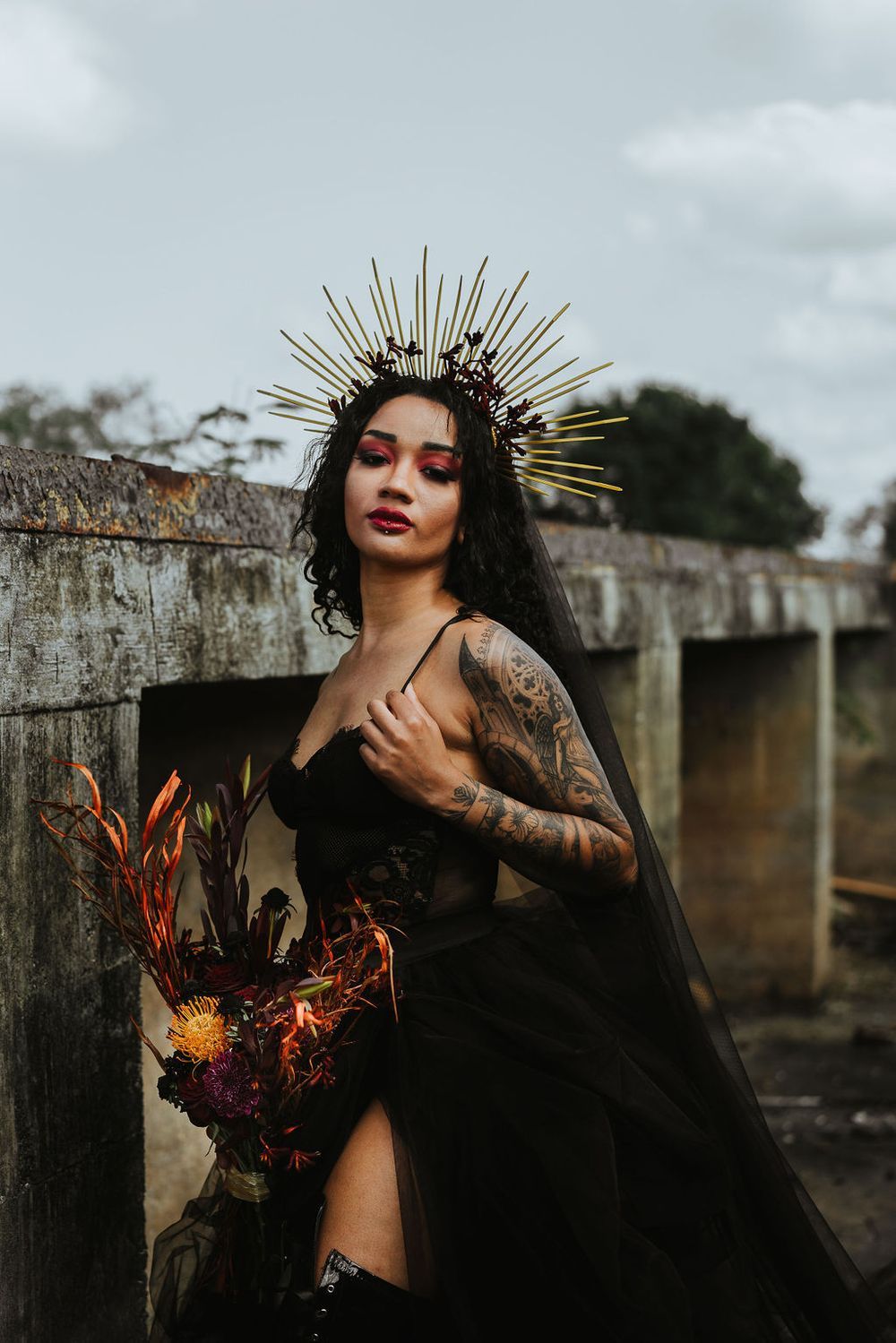 Gothic bride in black dress and golden crown headpiece poses with dark florals against stone wall.