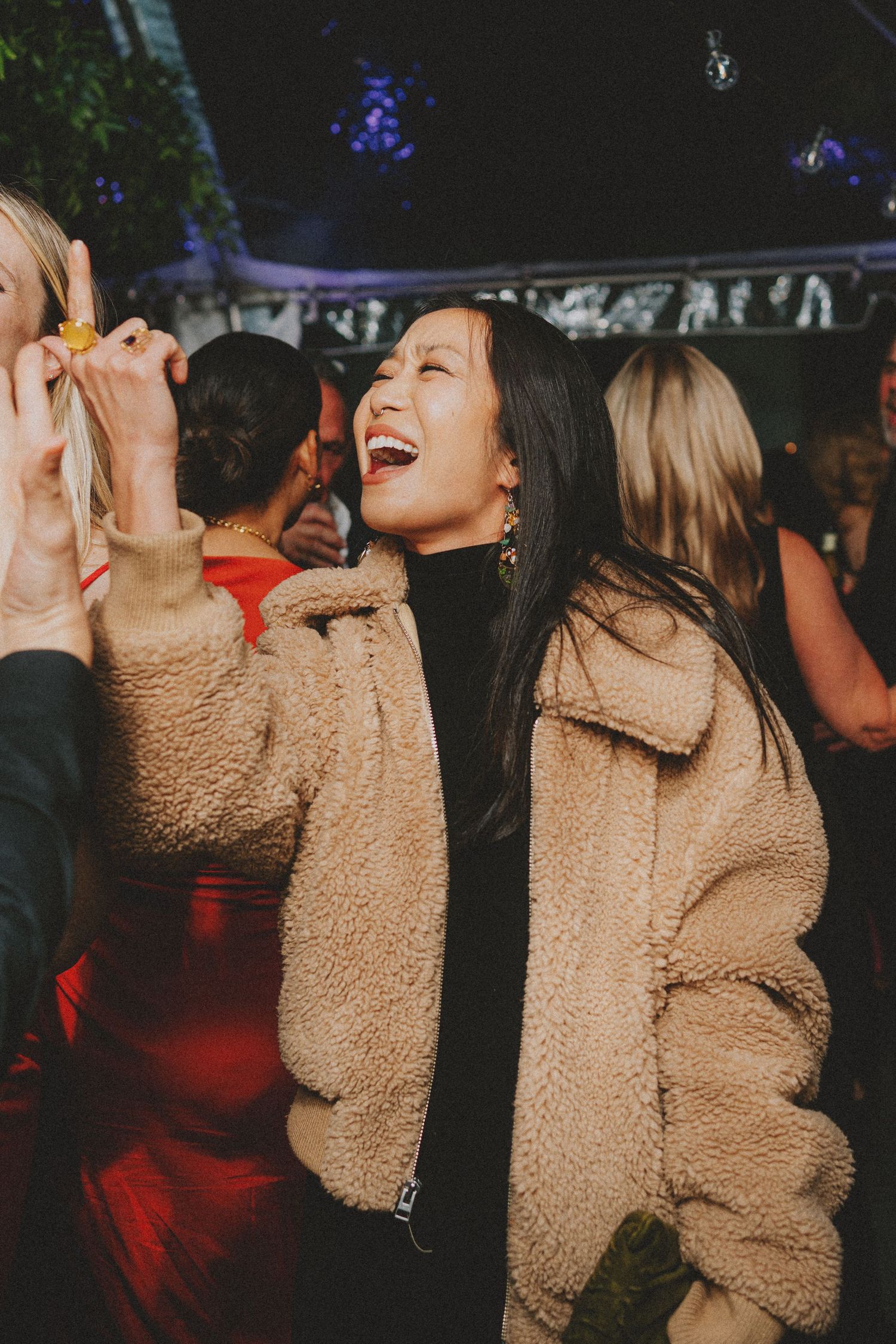 Woman in tan teddy coat laughing joyfully while dancing at outdoor evening wedding reception.
