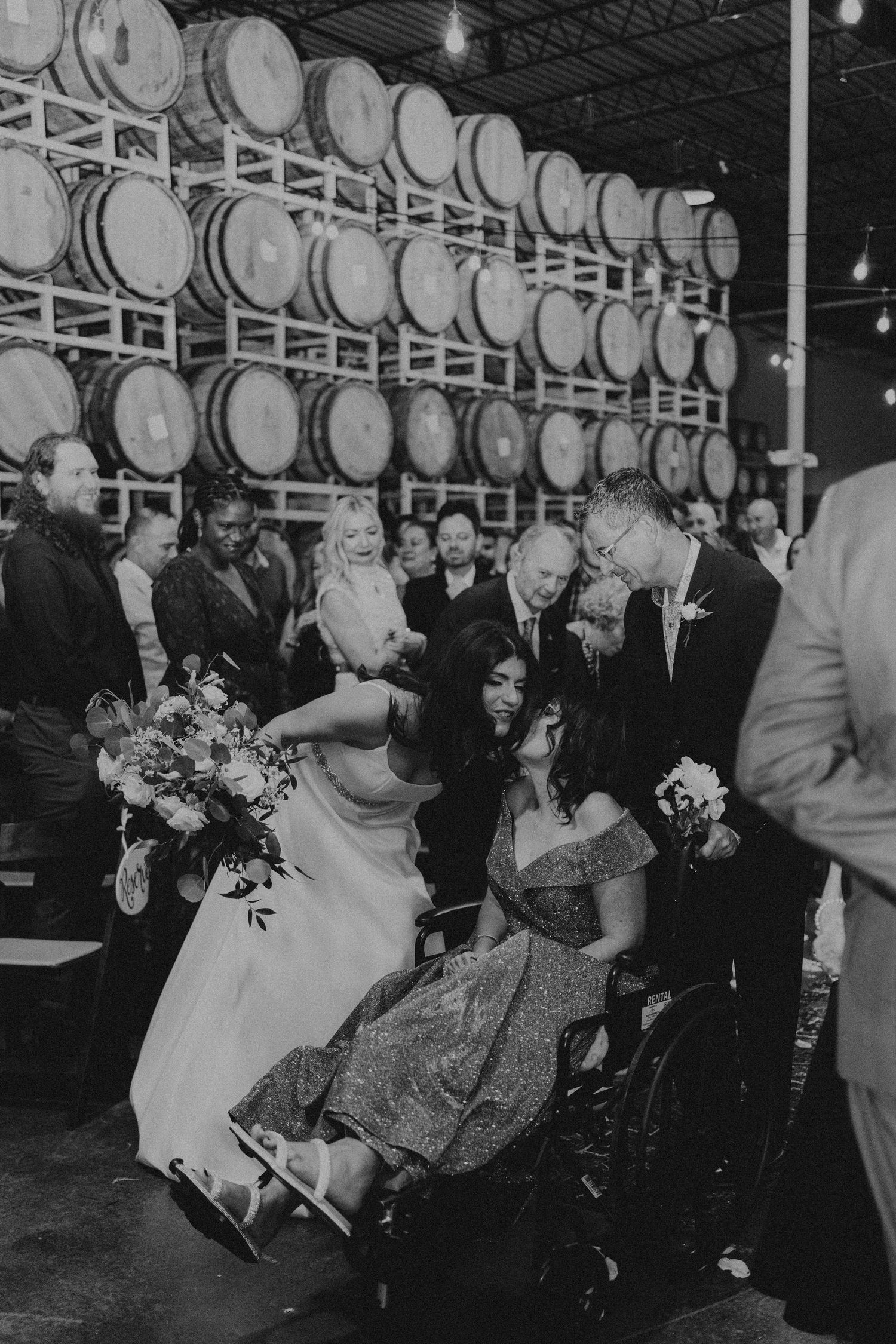 Black and white wedding photo at a winery with bride leaning to hug a guest in a wheelchair, barrels in background.