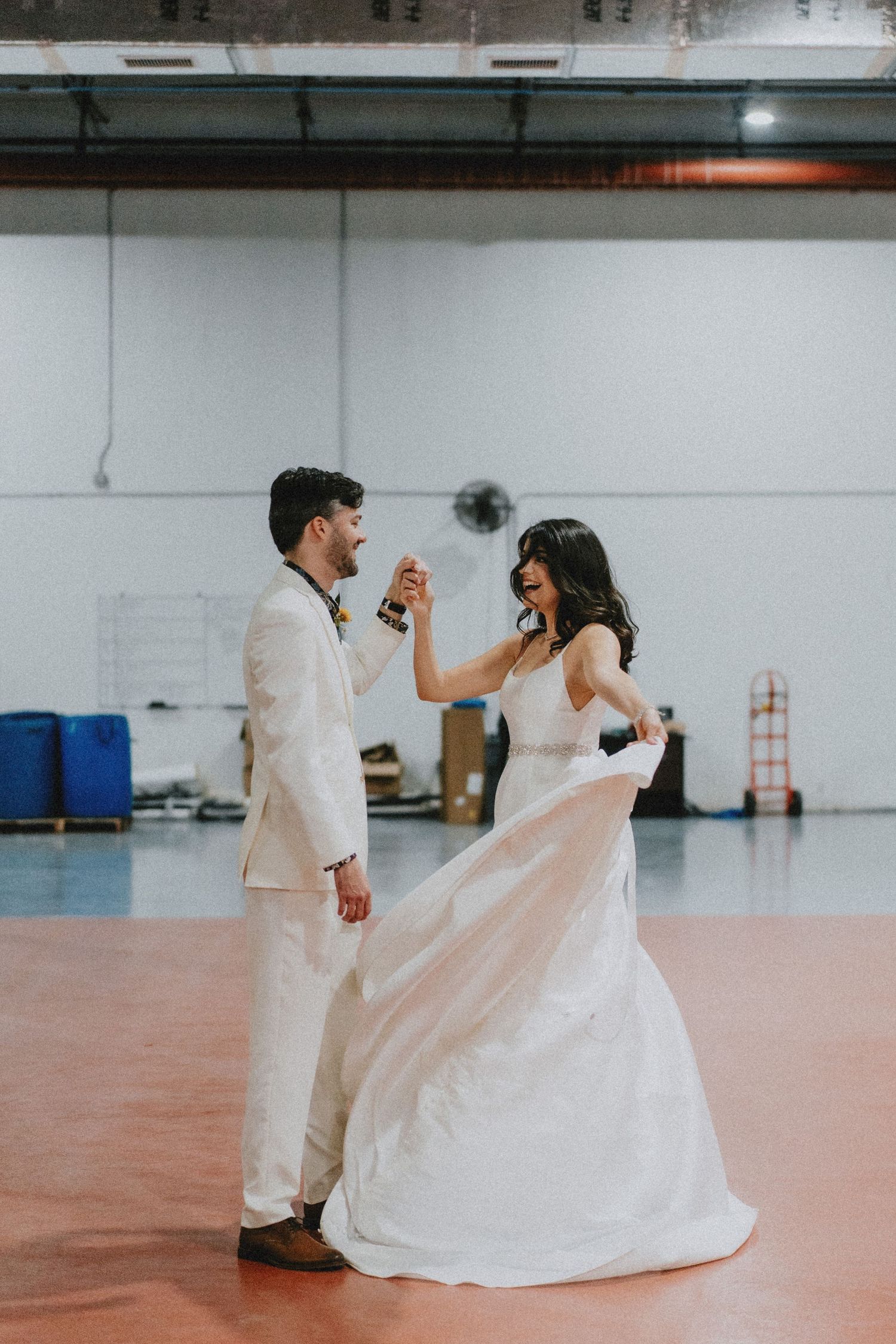 A bride in a flowing white gown twirls with a groom in a cream suit during their first dance in an industrial venue.