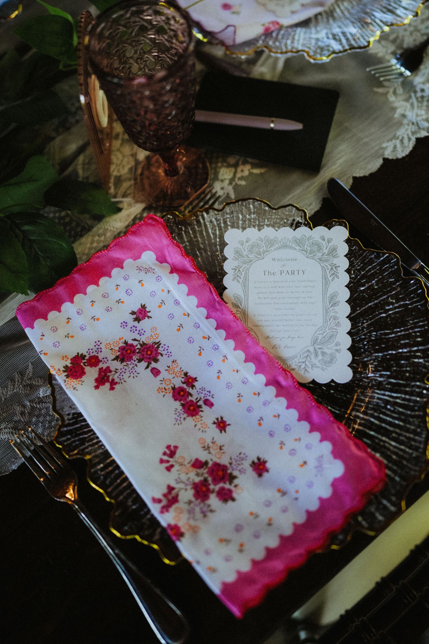 Elegant wedding table setting with floral embroidered napkin, scalloped menu card, and purple glassware on dark plate.