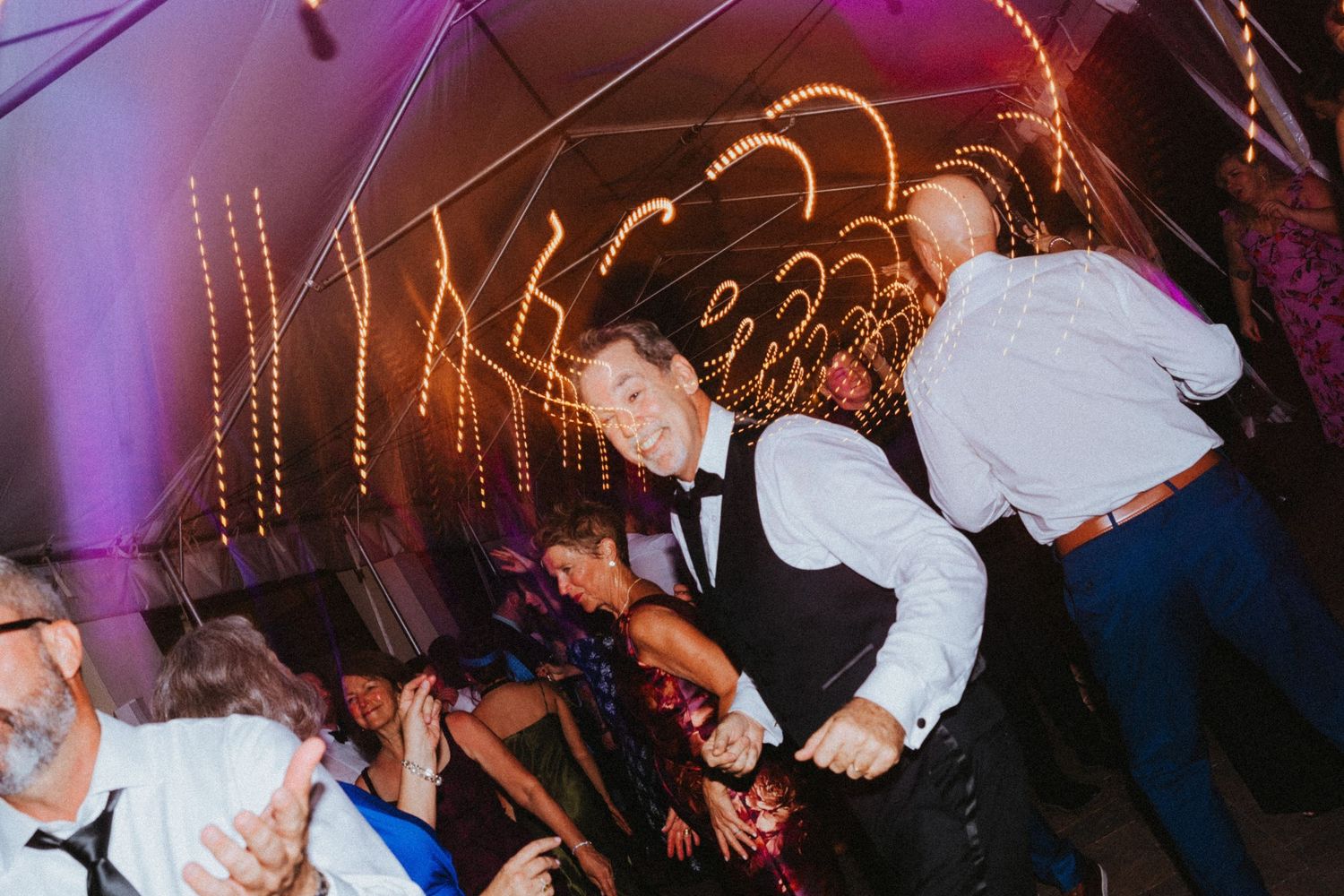 Energetic wedding guests dance enthusiastically under colorful lights at a lively indoor reception party.
