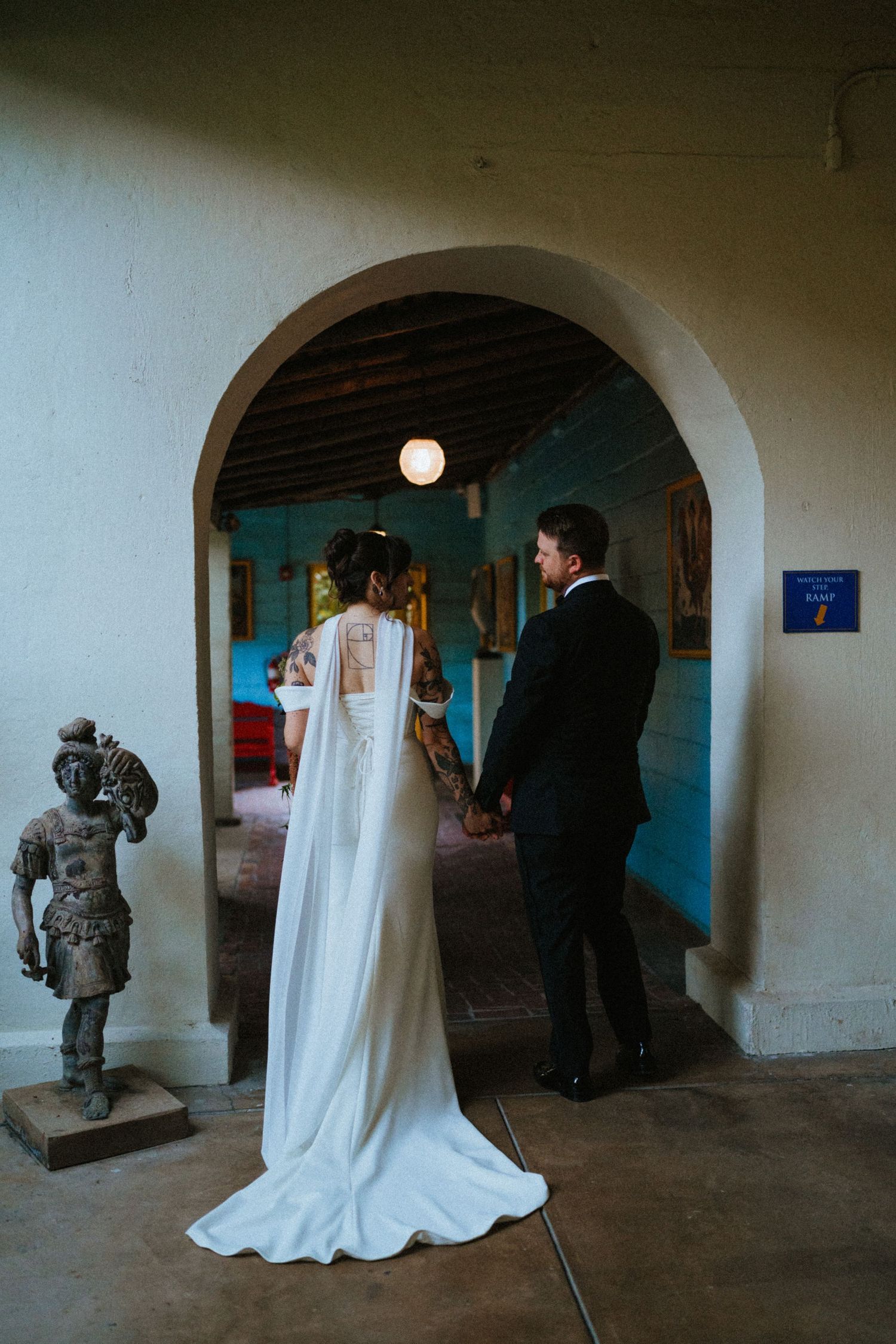 Bride and groom hold hands standing in an arched doorway of a rustic venue with a bronze statue nearby.