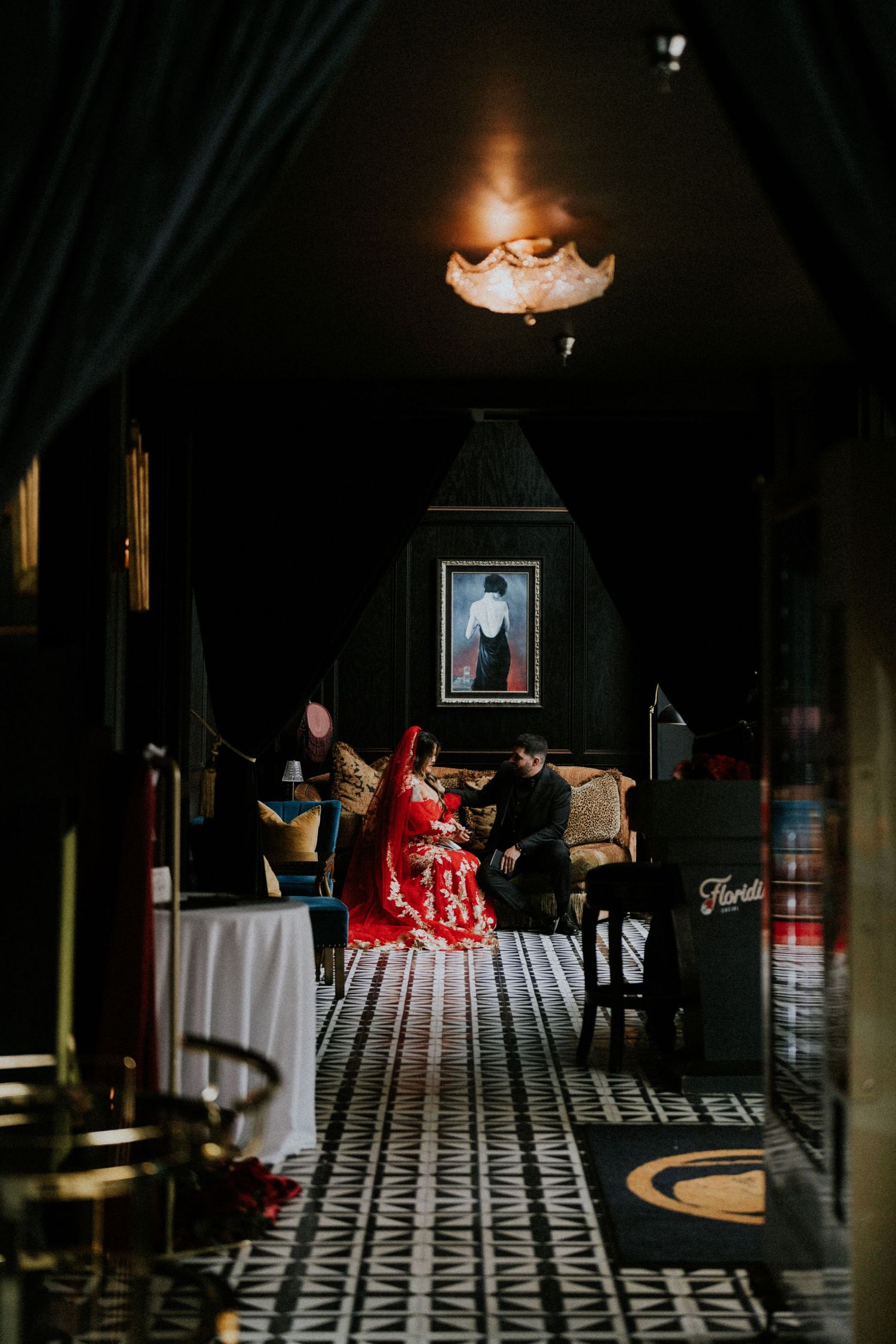Couple in traditional red wedding attire seated in an elegant dark-walled room with patterned floors and warm lighting.