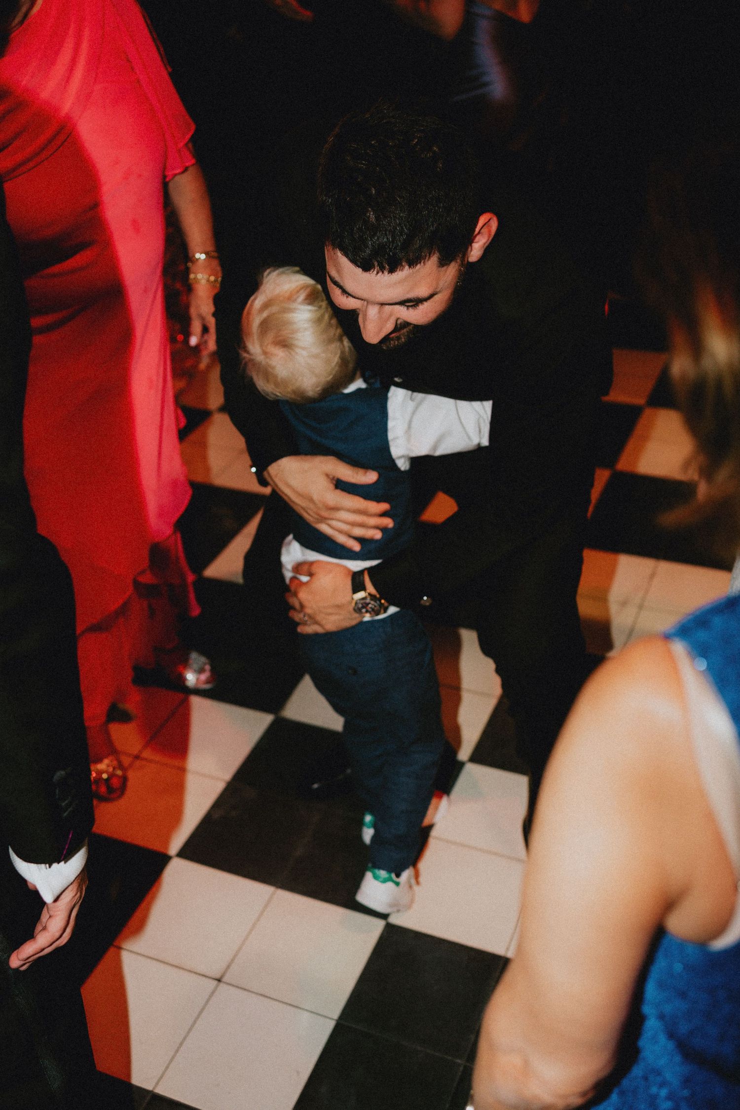 Man in black suit dancing with a toddler boy on a checkered dance floor at a formal event.