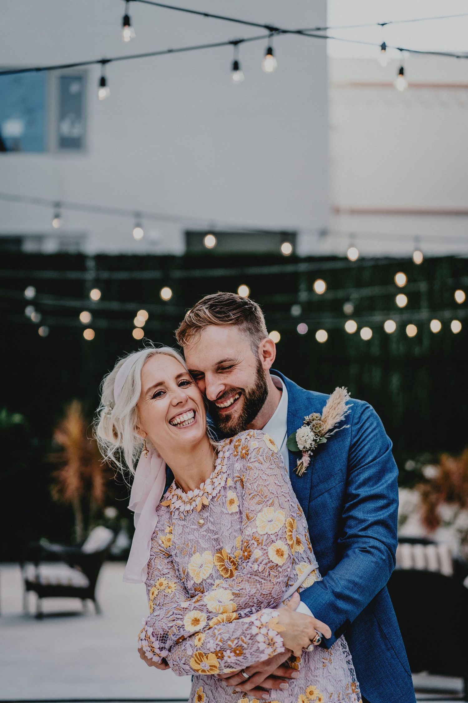 A joyful couple embraces outdoors at their wedding, with string lights glowing above them at dusk.
