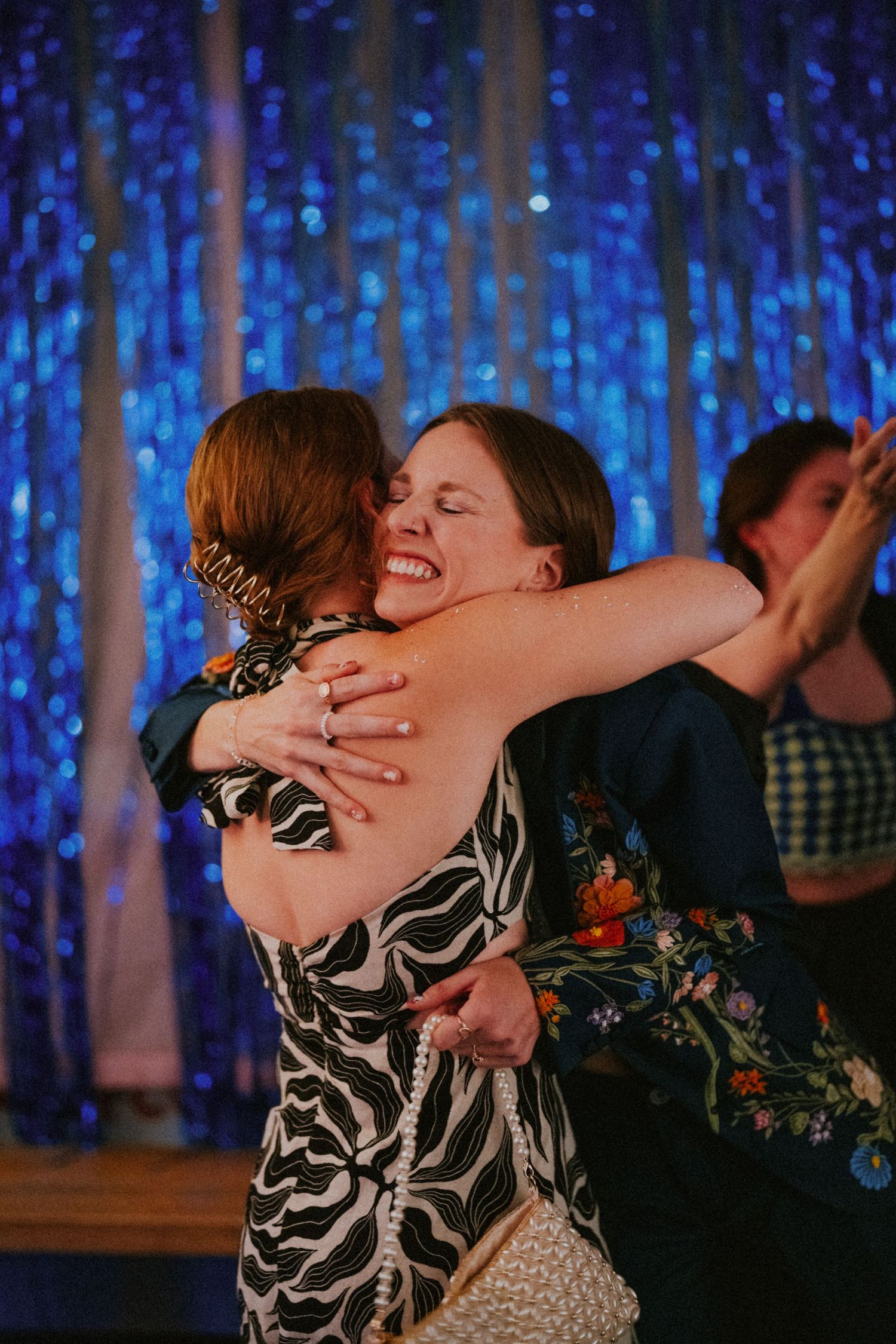 Two women share a joyful hug on a dance floor in front of a sparkling blue sequin backdrop.